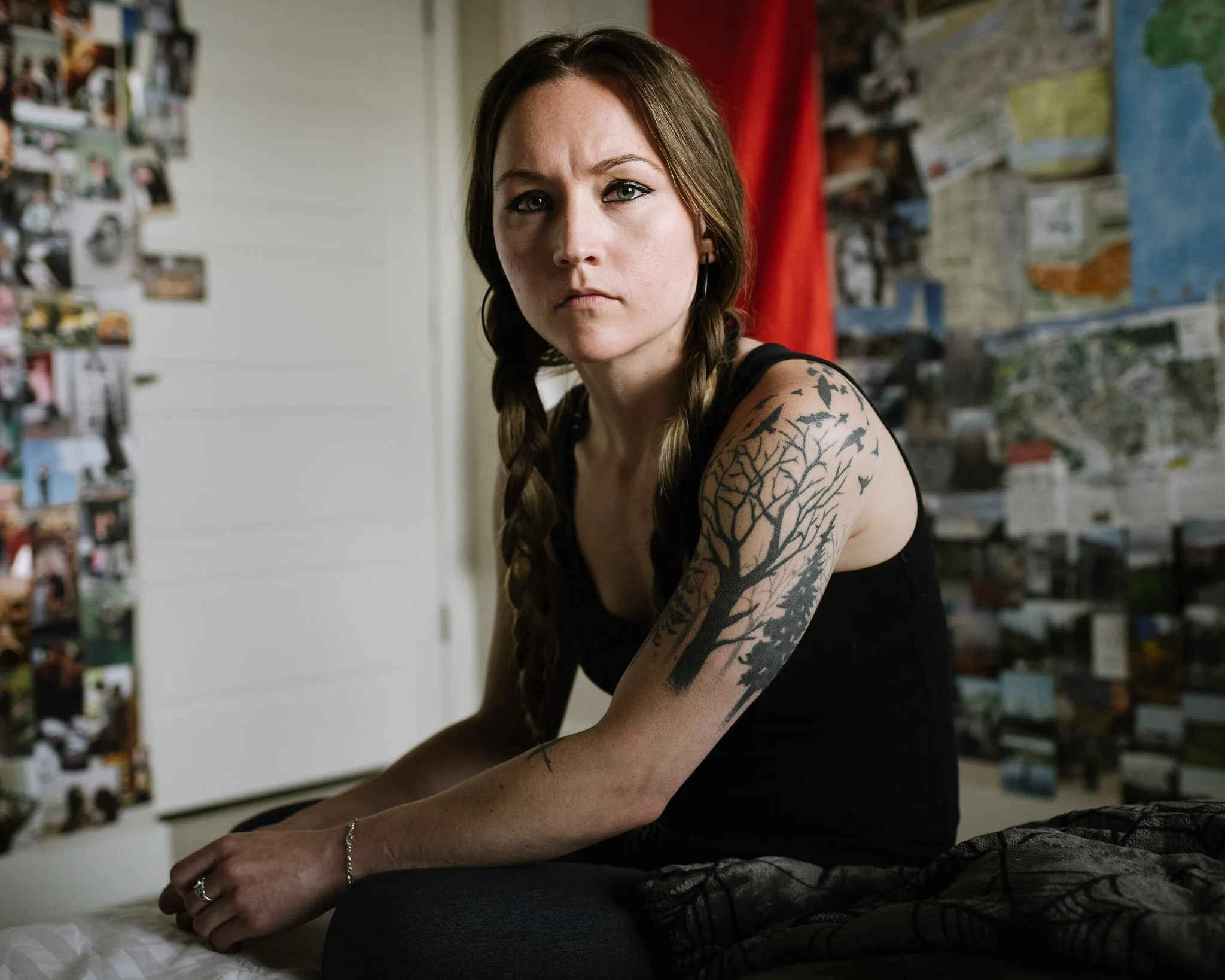 White woman brown hair in long braids, intense stare,  sitting on the edge of her bed, the walls behind her coverd in small photographs and a red drape. She wears a black tanktop and her her arm has a prominant tattoo of birds and a forboding tree.