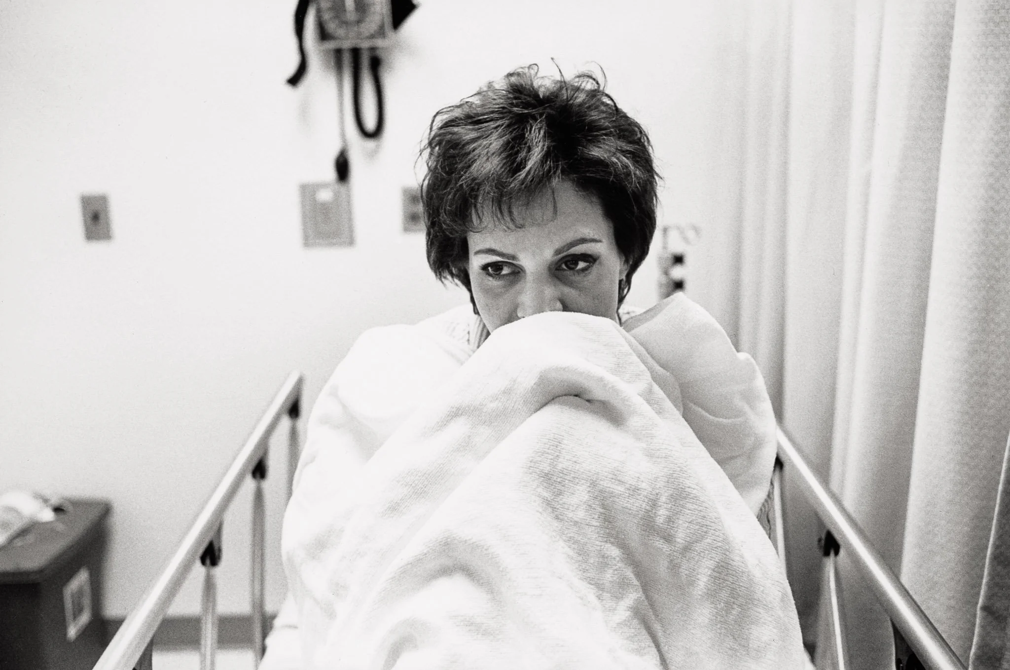 A woman in a  surgical preparation room huddles under her blanket, with nervous trepidation before her mastectomy.