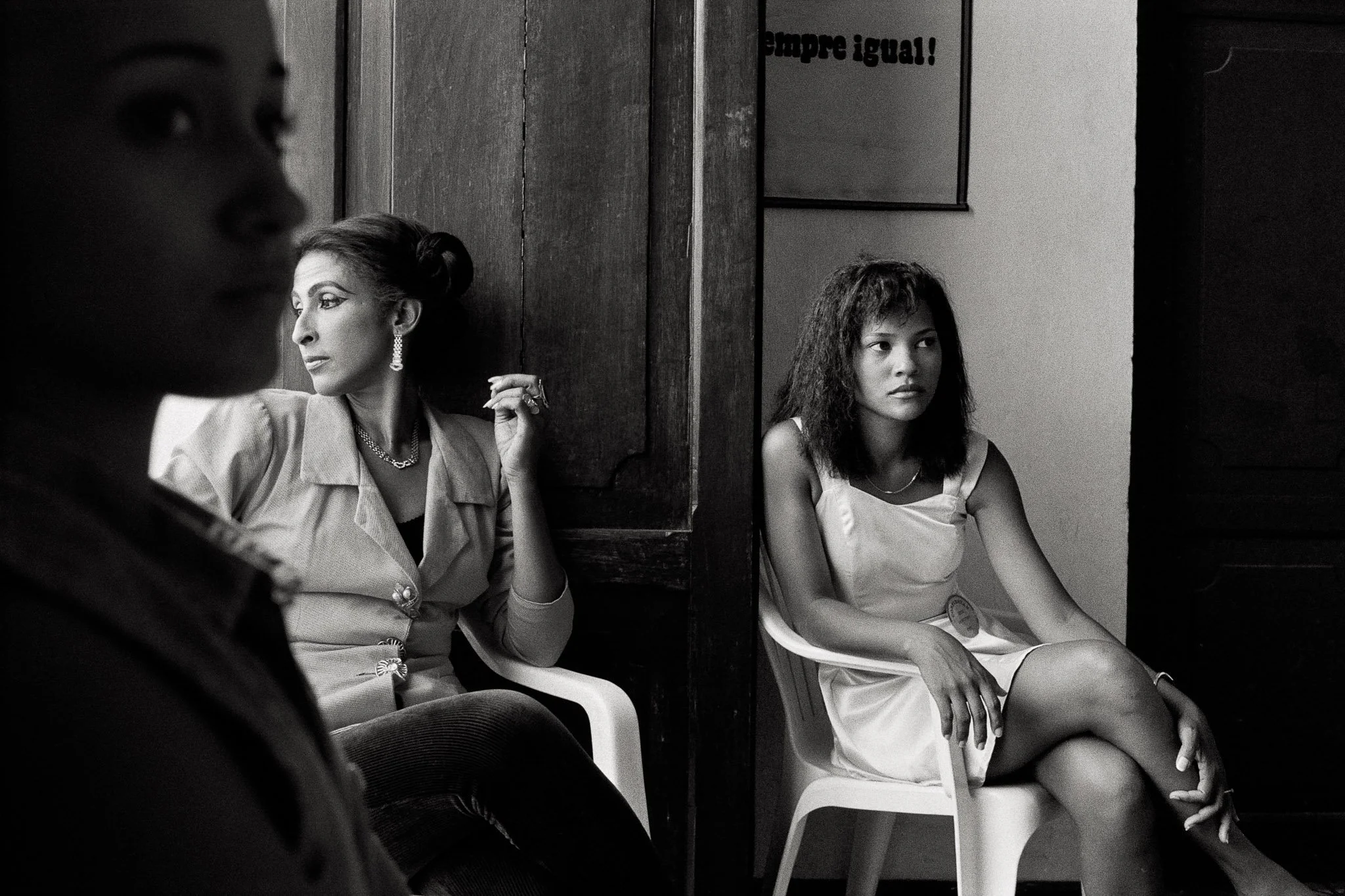 Black and white image of three queens of three generations in moody light, each in their own worlds, connotations of cultural continuity. A sign on the wall says "Siempre Igual".