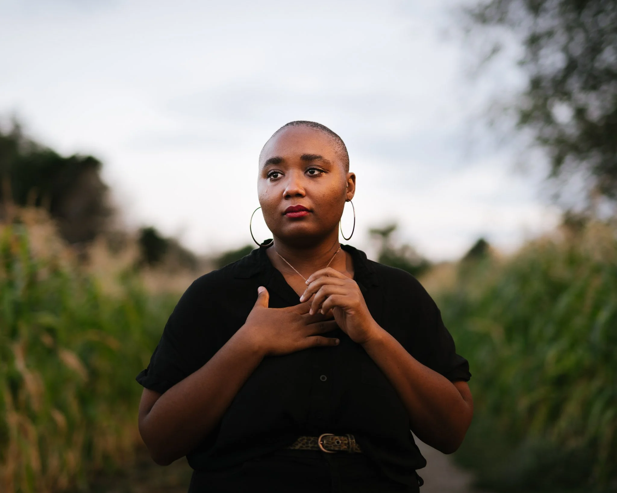 Beautiful young black woman with cropped hair and large earrings in a cornfield at dusk, her hand toucher her chest with emotion as a tear falls from her eye.