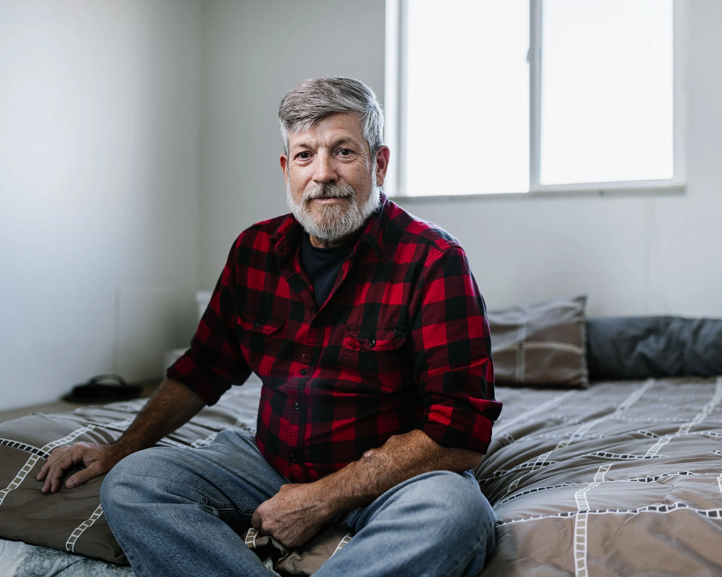 Portrait of a handsome and formerly homeless veteran in a red plaid shirt sitting on the corner of his bed, soft bright light coming through the window.