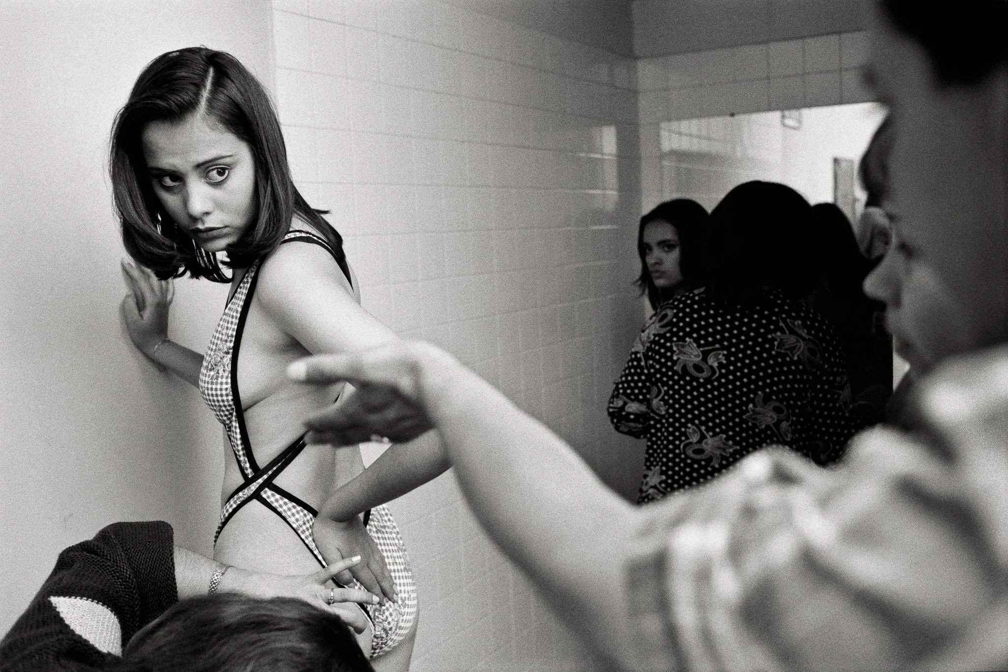 A Colombian beauty pageant contestant in her swimsuit in a changing room. She is thin, but a makeup artist is trying to disguise very subtle signs of cellulite on her buttocks.  Her coach makes a critical gesture and the girl looks concerned.