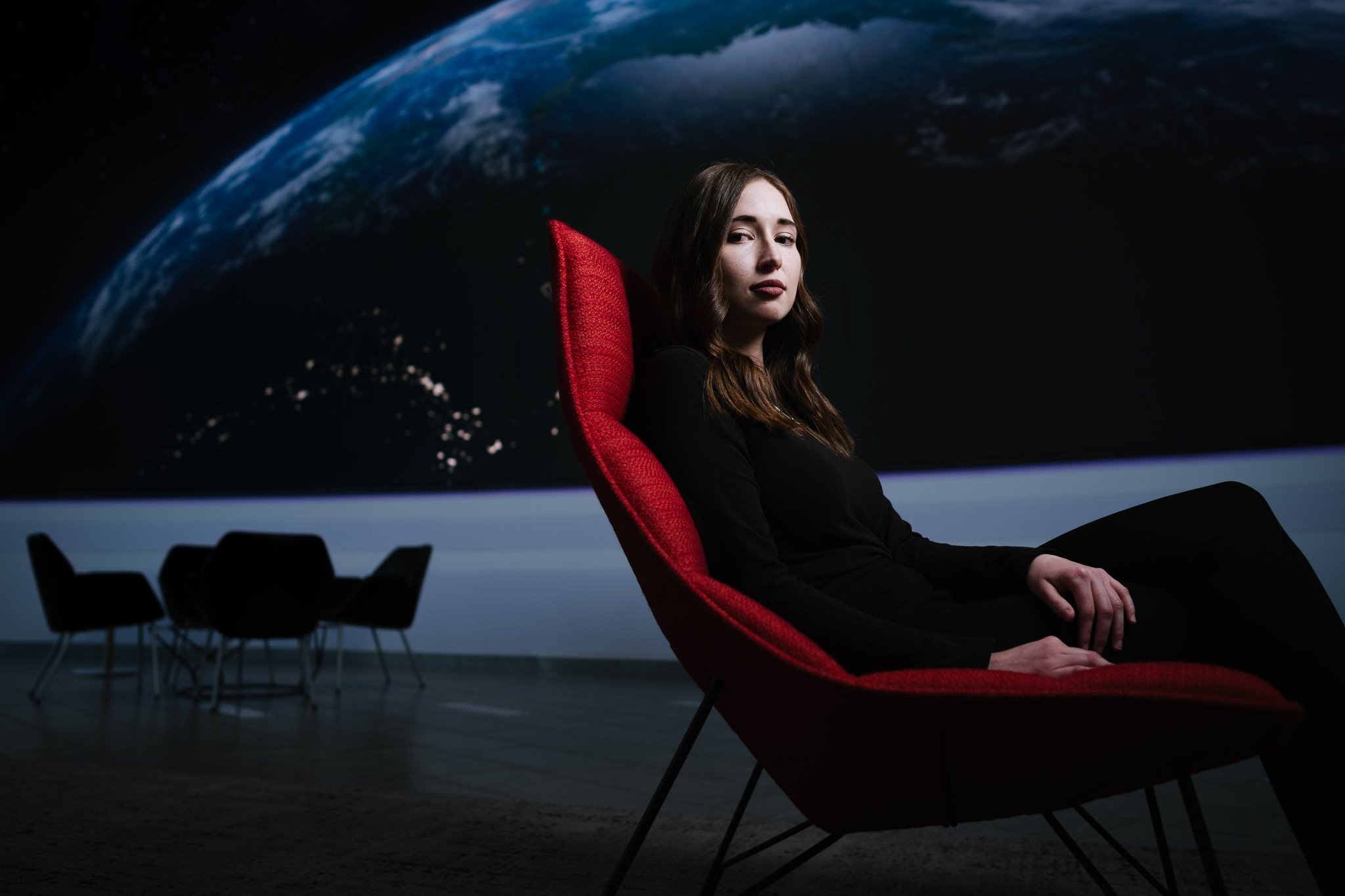 Dramatically lit portrait of astronautical engineer Renee Eastburn in a red chair in a dark room, a mammoth screen of the earth from low orbit behind her. The photo is black, red, blue and the pale color of her skin.