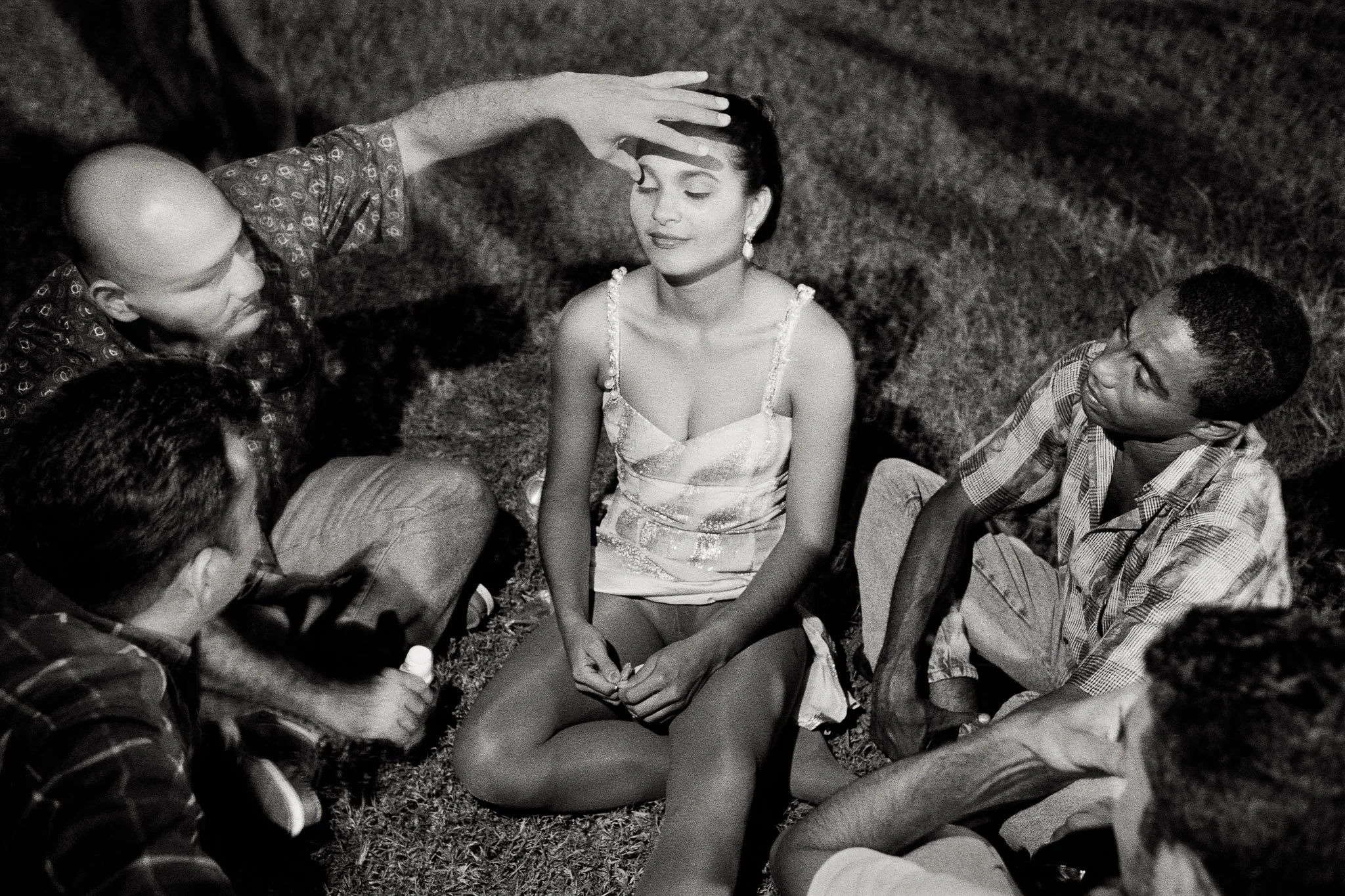 A beauty queen sits in the grass after a contest, surrounded by four older men. Her dress is pulled up to her waist, her thong visible. A man with a bottle of hard liquor puts his hand on her forehead, her eyes closed.