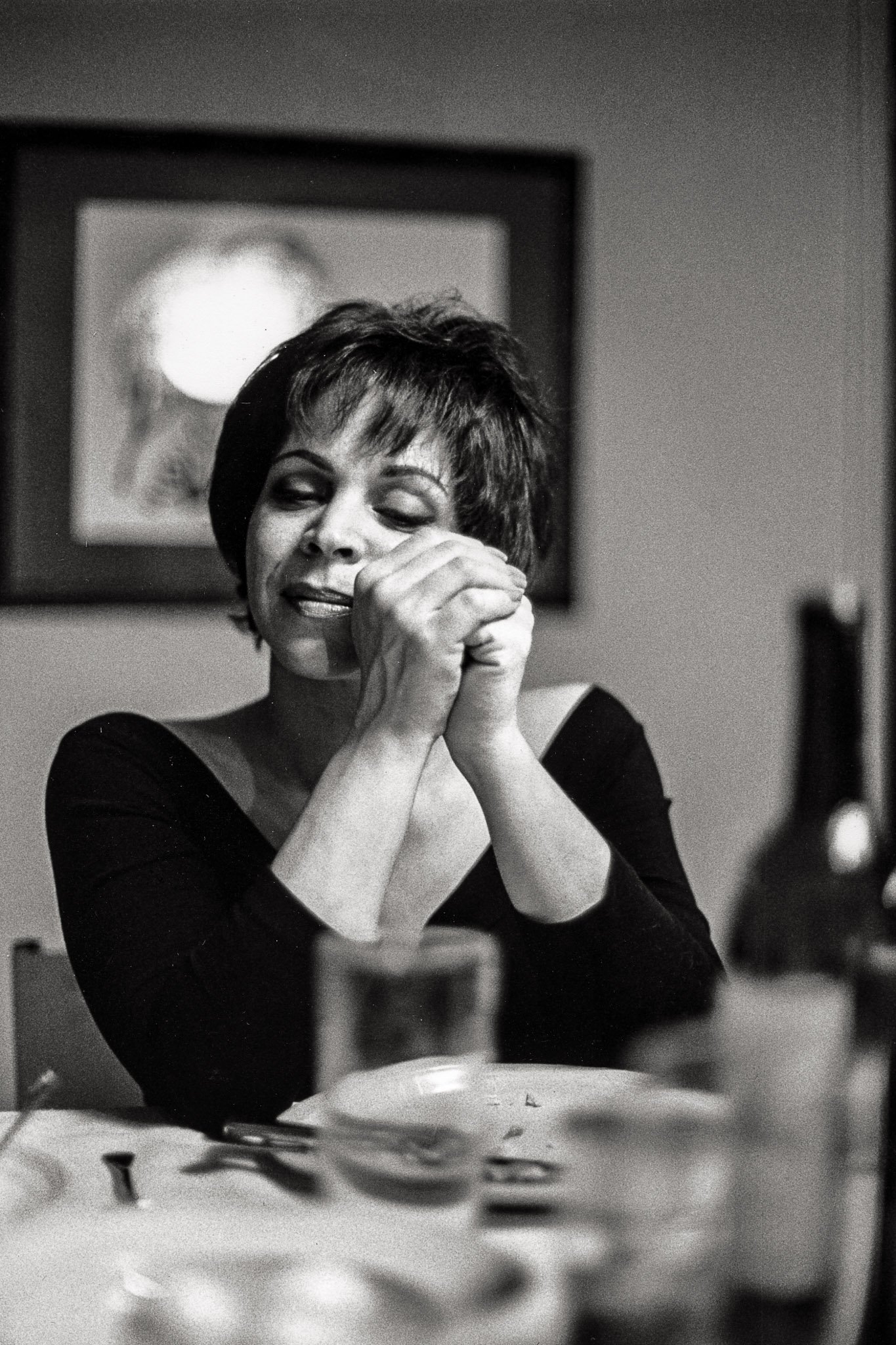A breast cancer survivor sits at her table during Passover, hands folded in front of her face, head tilted, eyes closed, smiling.