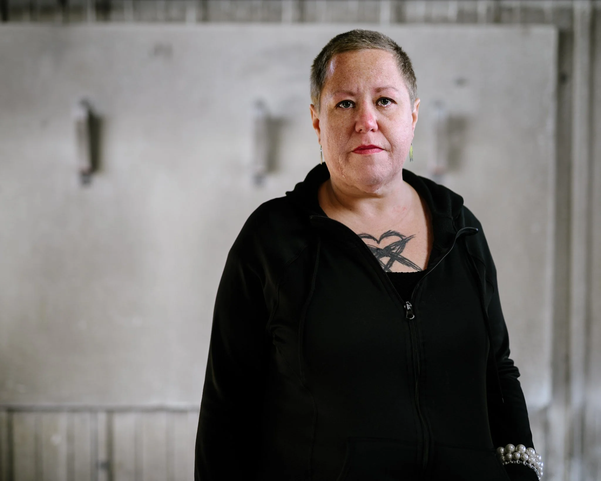 A white woman with closely-cropped gray hair, a black hoodie, a pearl bracelet and a tattoo on her chest of a large heart with an X through it. Tears well in her eyes as she is lost in thought. The background is non-descript, gray, industrial.