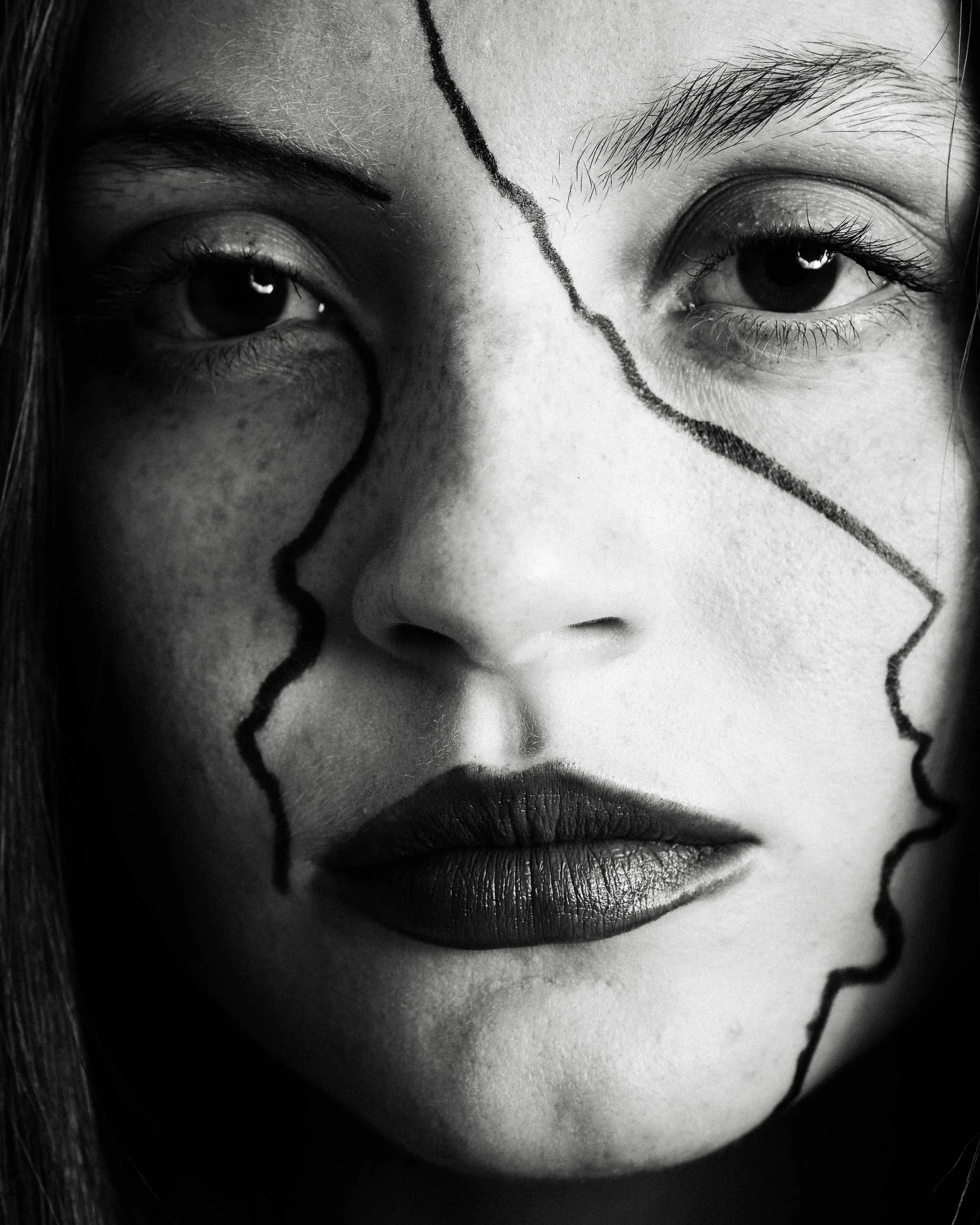 Graphic black and white close up portrait of a white woman with black lipstick and eyeliner in jagged lines across her face as if wearing a mask that is cracking.