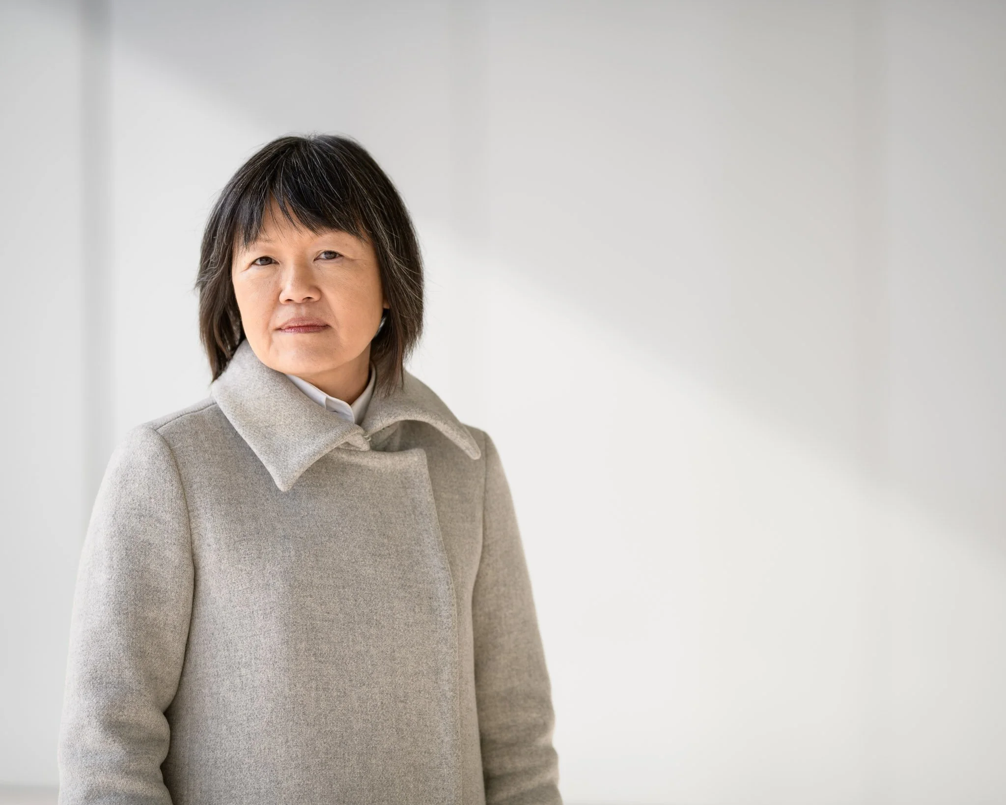 High key minimalist portrait of architect Pamela Sams, her off-white coat blending with the wall behind her, mirroring the minimalism of her design.