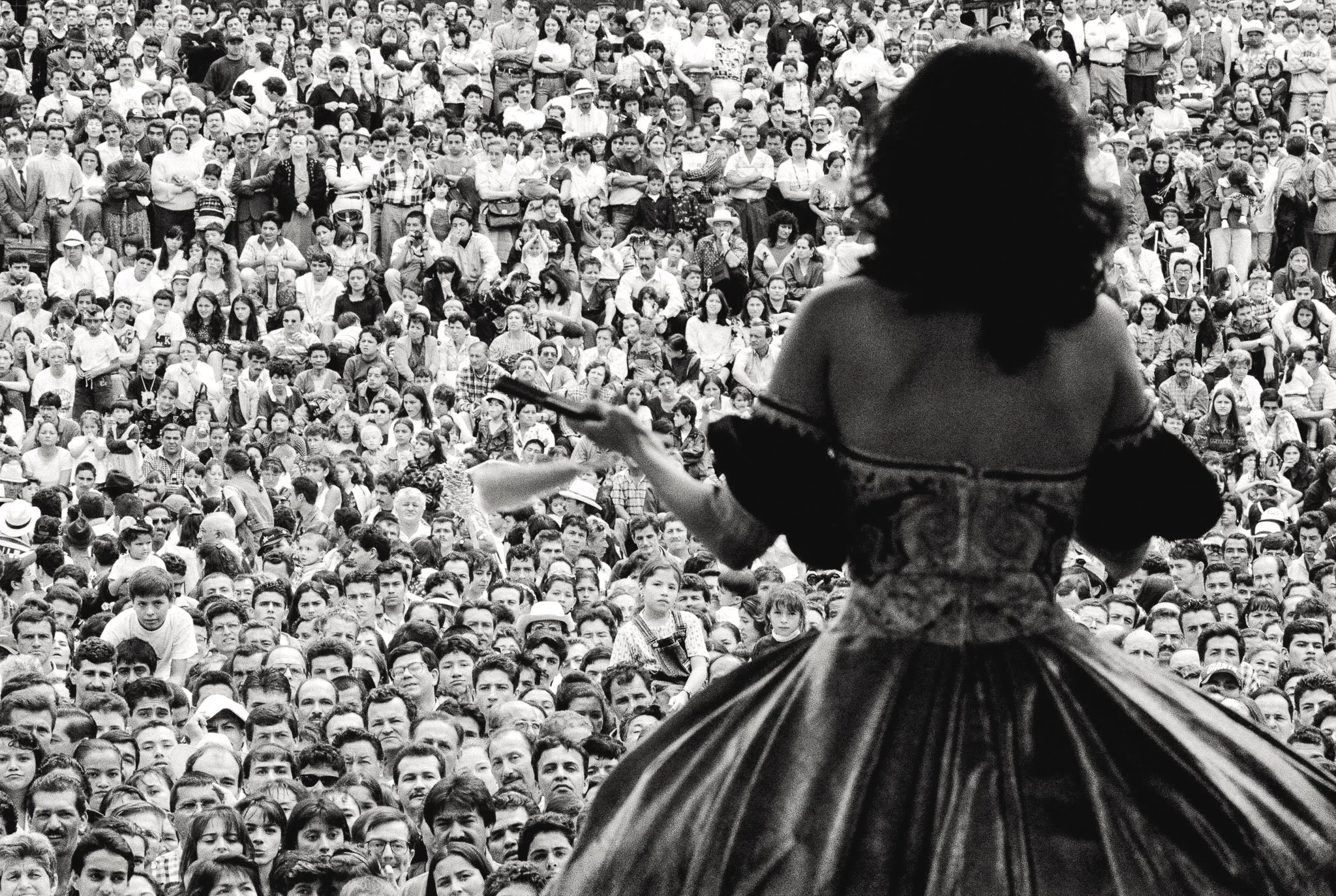 Image of a beauty queen in a ballgown before a sea of thousands of faces. She is viewed from behind as her viewers' faces fill the rest of frame.