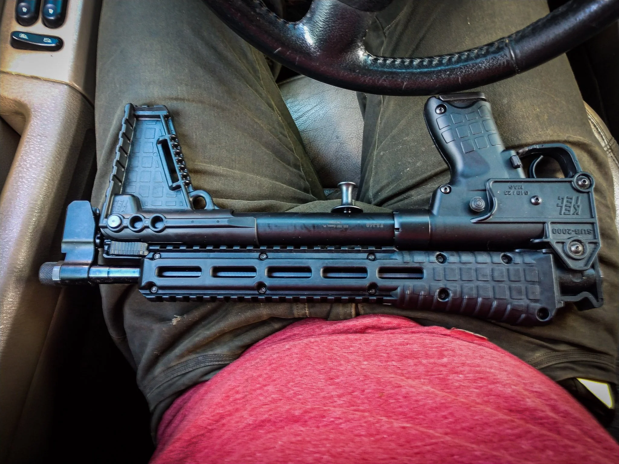 Photograph of a folded assault rifle on a man's lap as he sits in his car, the barrel folded back against the stock. Brand is KelTel. He is proud that he can slip his assault rifle into a small backpack and pass unnoticed.