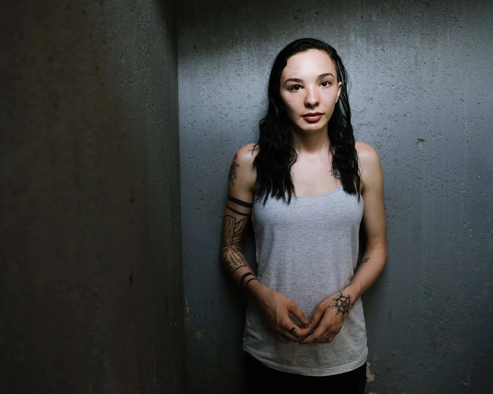 A cold-tone image of a young woman in a concrete basement starewell, dim lighting, her pale skin glowing in the darkness. There are subtle signs of facial surgery. She wears a gray camisole, tattoos cover her arms, including a pentagram on her hand.