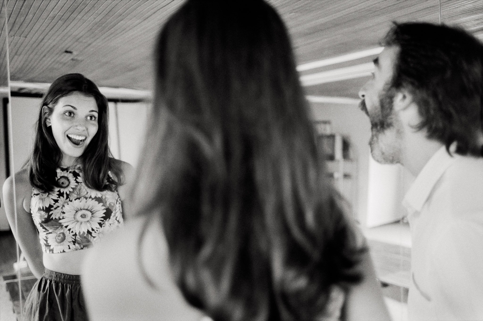 A Colombian beauty pageant contestant in workout clothing stands in front of a mirror with her coach and practices looking pleasantly surprised.