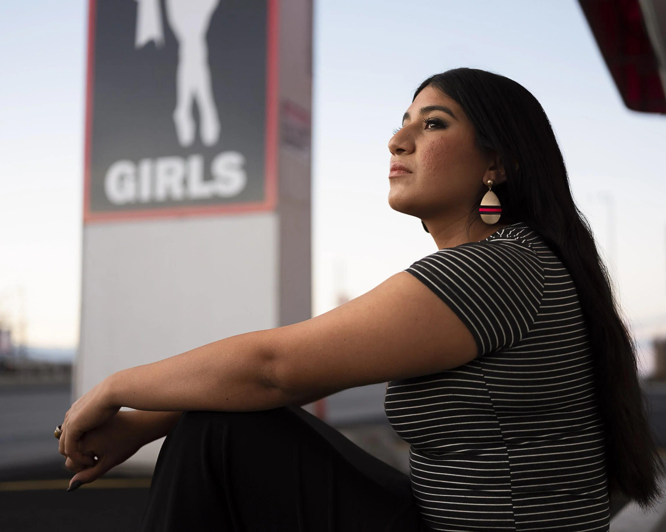 Latina in a tightly-fitted shirt sits in the parking lot of a strip club at sunset, looking into the distance, the  club signage in the background behind her. Image dominated by the colors black, white and red.