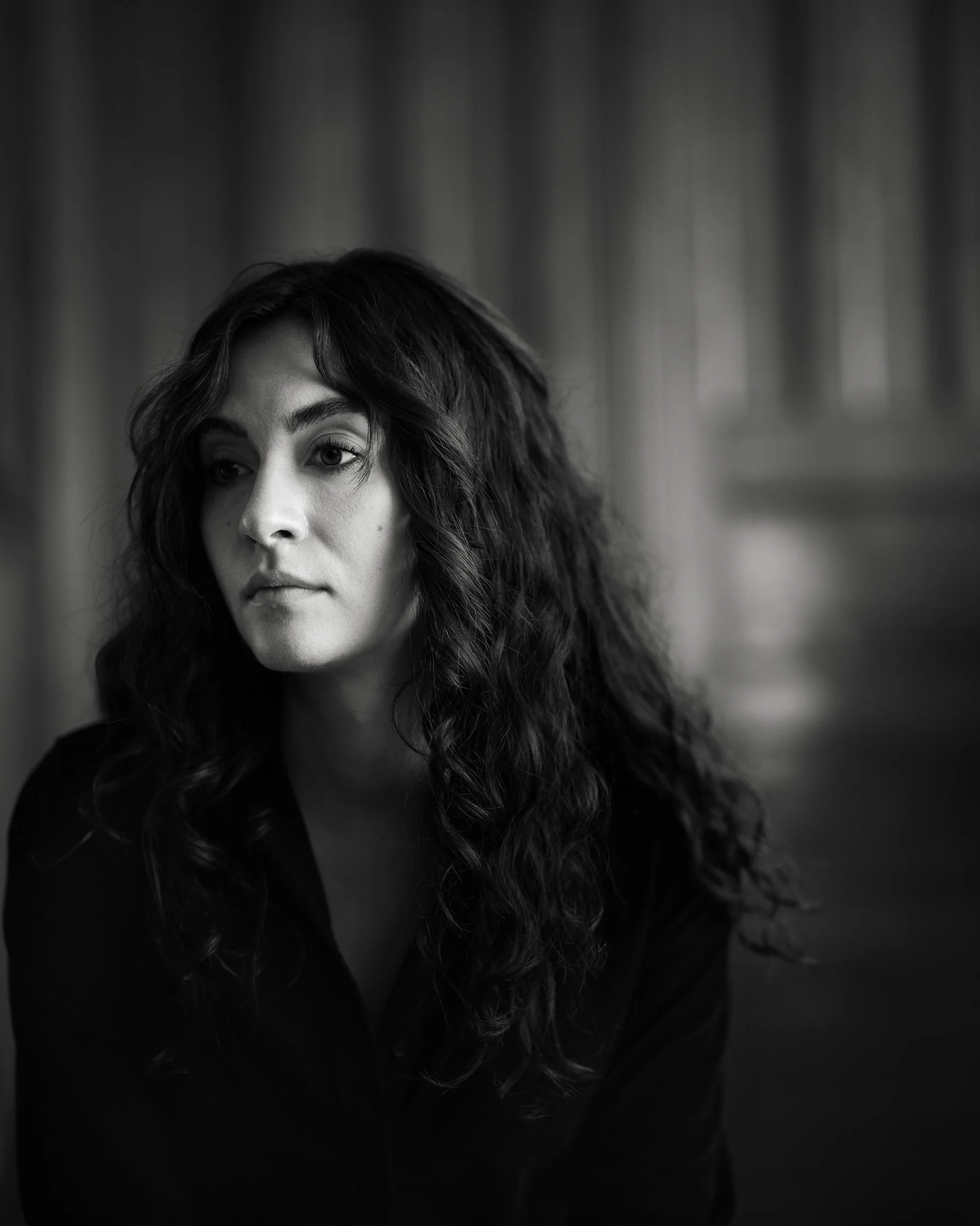 Black and white moody portrait of woman with long black curly hair in a dim room, shallow focus, head turned away, contemplative expression.