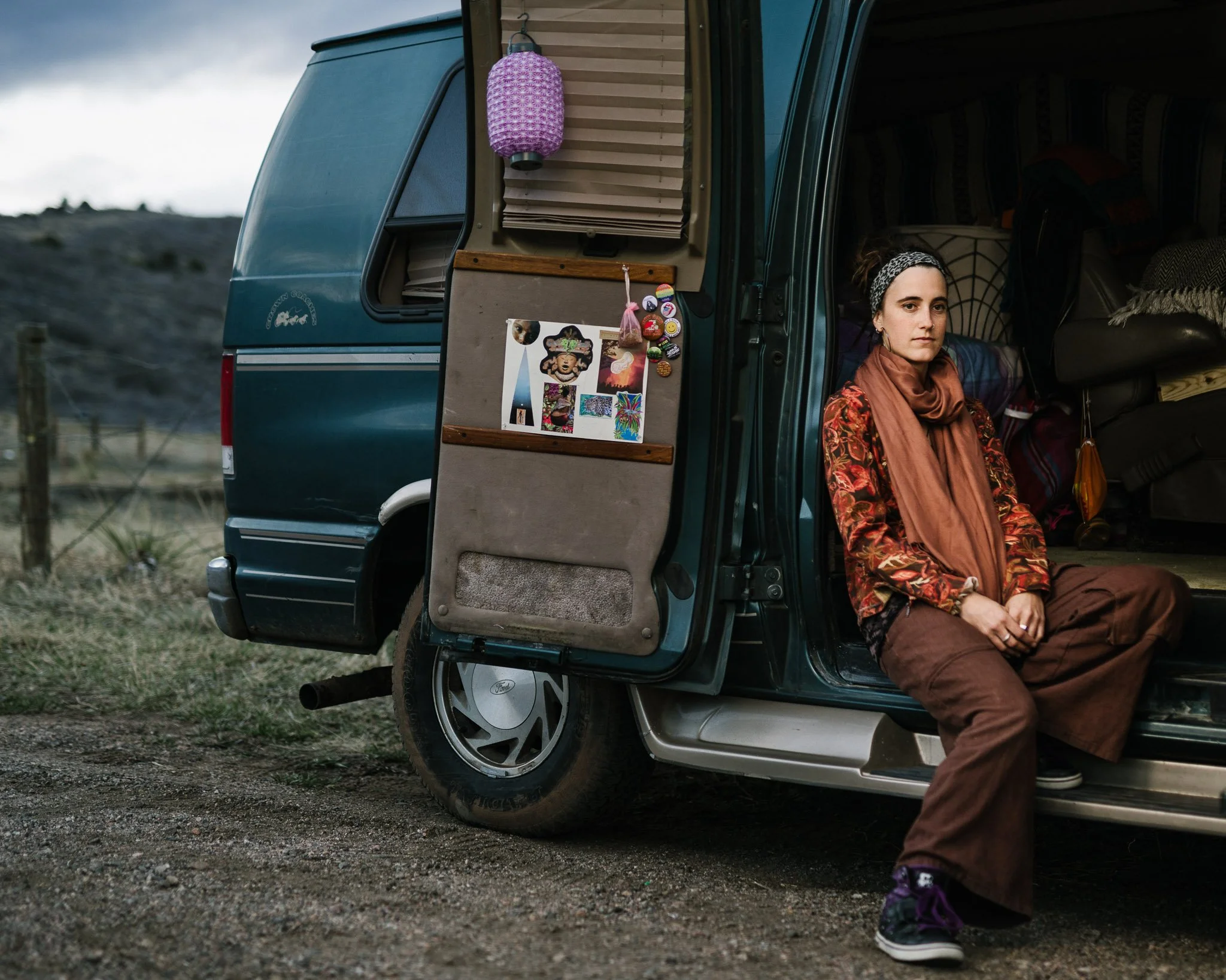 A Spanish woman sits in the side of the 70s van she lives in, parked in high desert in Colorado. She wears a headband and long flowing clothing and a large scarf, buttons and a lantern hanging on the van's open door.