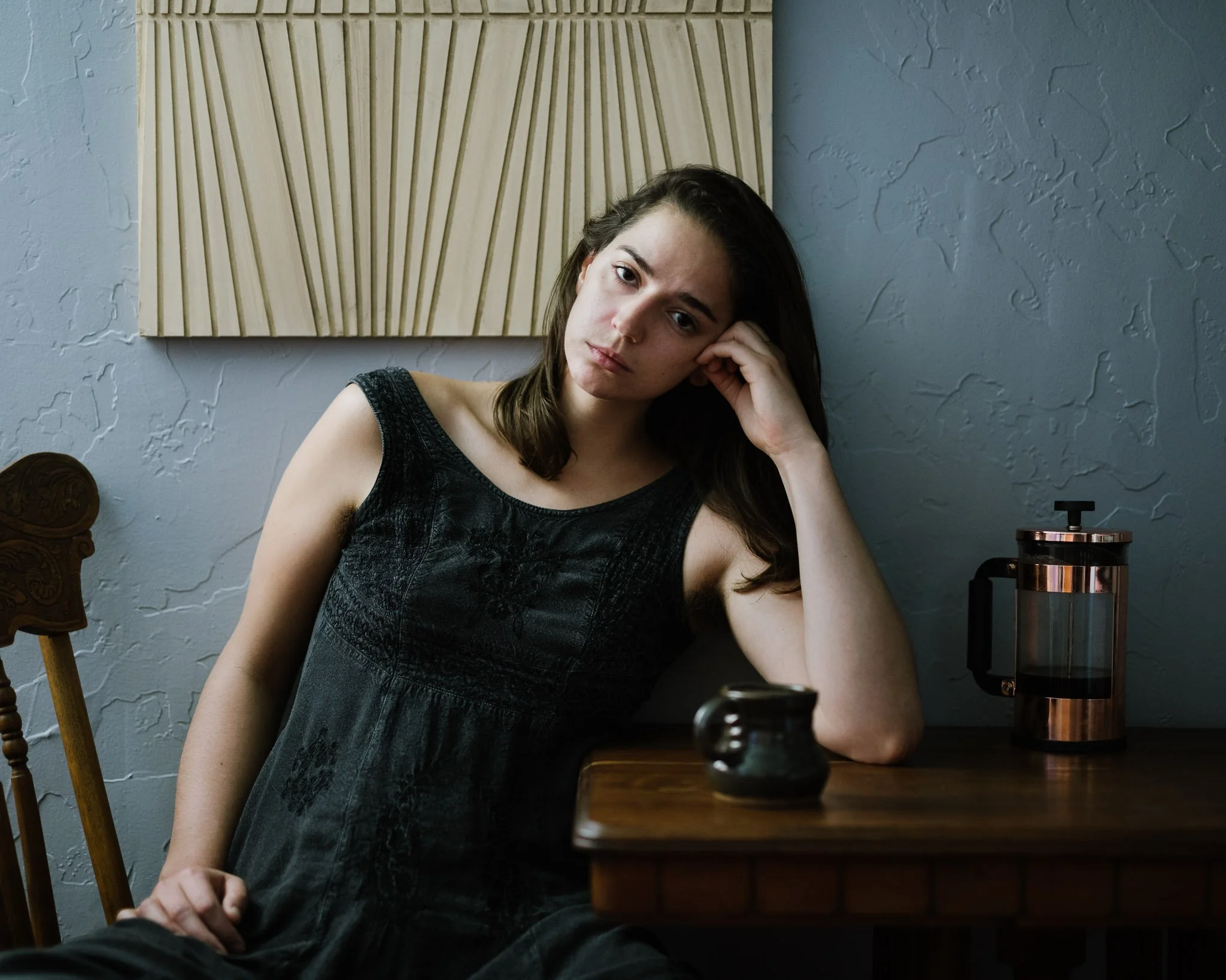 Woman in her 20s with pale skin, brown hair, and a black dress sitting at her small table against a blue wall, head propped on one hand, her coffee cup on the table, looking at viewer with a sad expression.