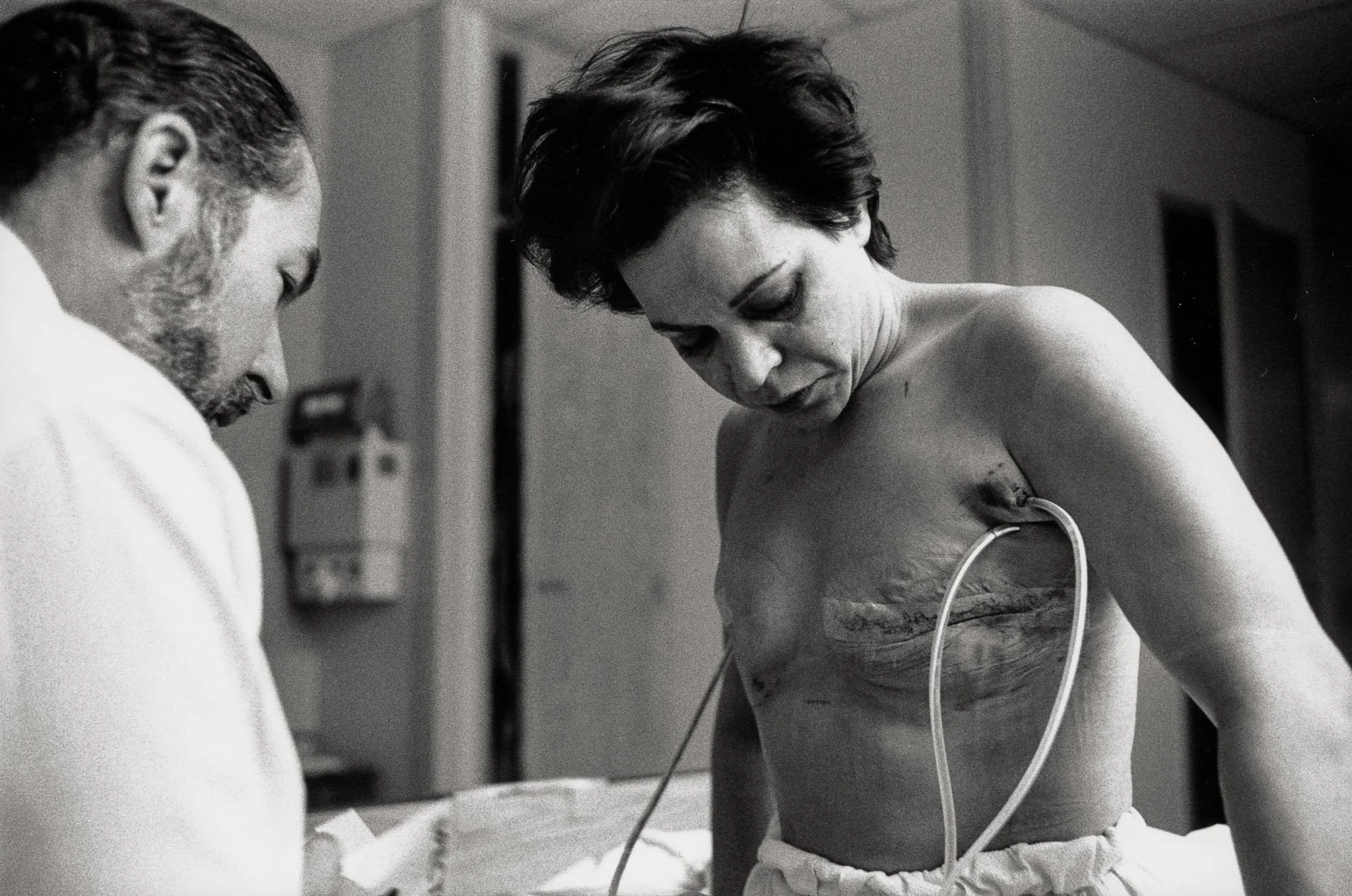 A woman sits up in her hospital bed the morning after her mastectomy, viewing the scars on her chest for the first time. She has dark circles under her eyes, her wounds are raw, drainage tubes come from her pectoral area.