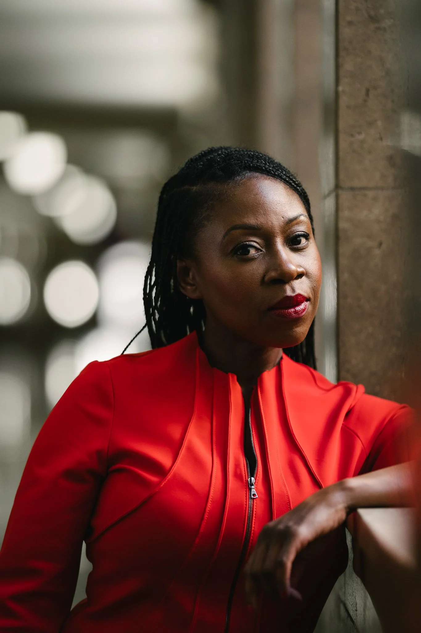 Dramatic portrait of Denver COO Janel Forde in a structured bright red dress with black zipper, her elbow resting on a window well in City Hall.