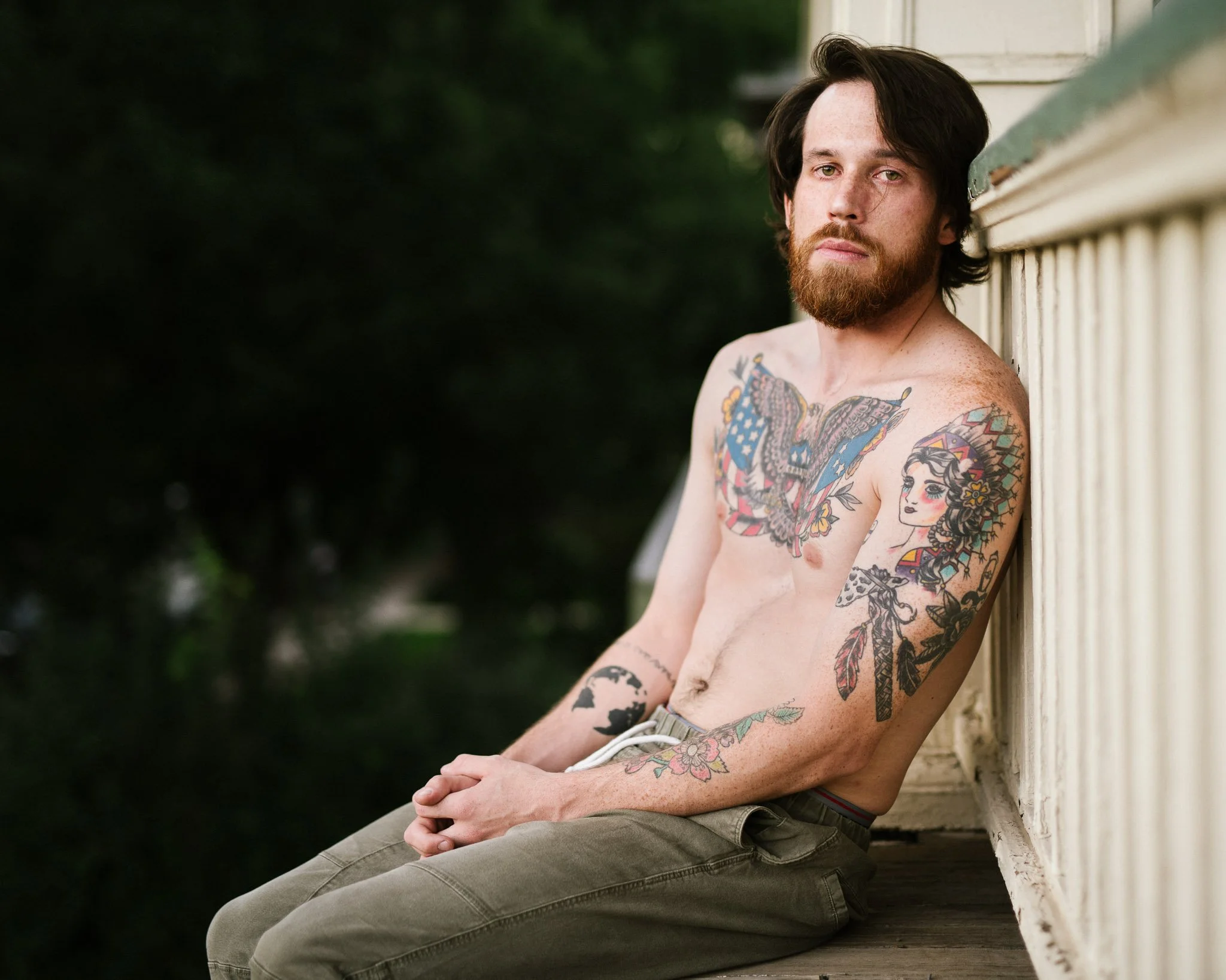 A white army veteran with long brown hair and a full beard sits on the edge of a balcony. His shirt is off, revealing large tattoos with patriotic and Native American images.