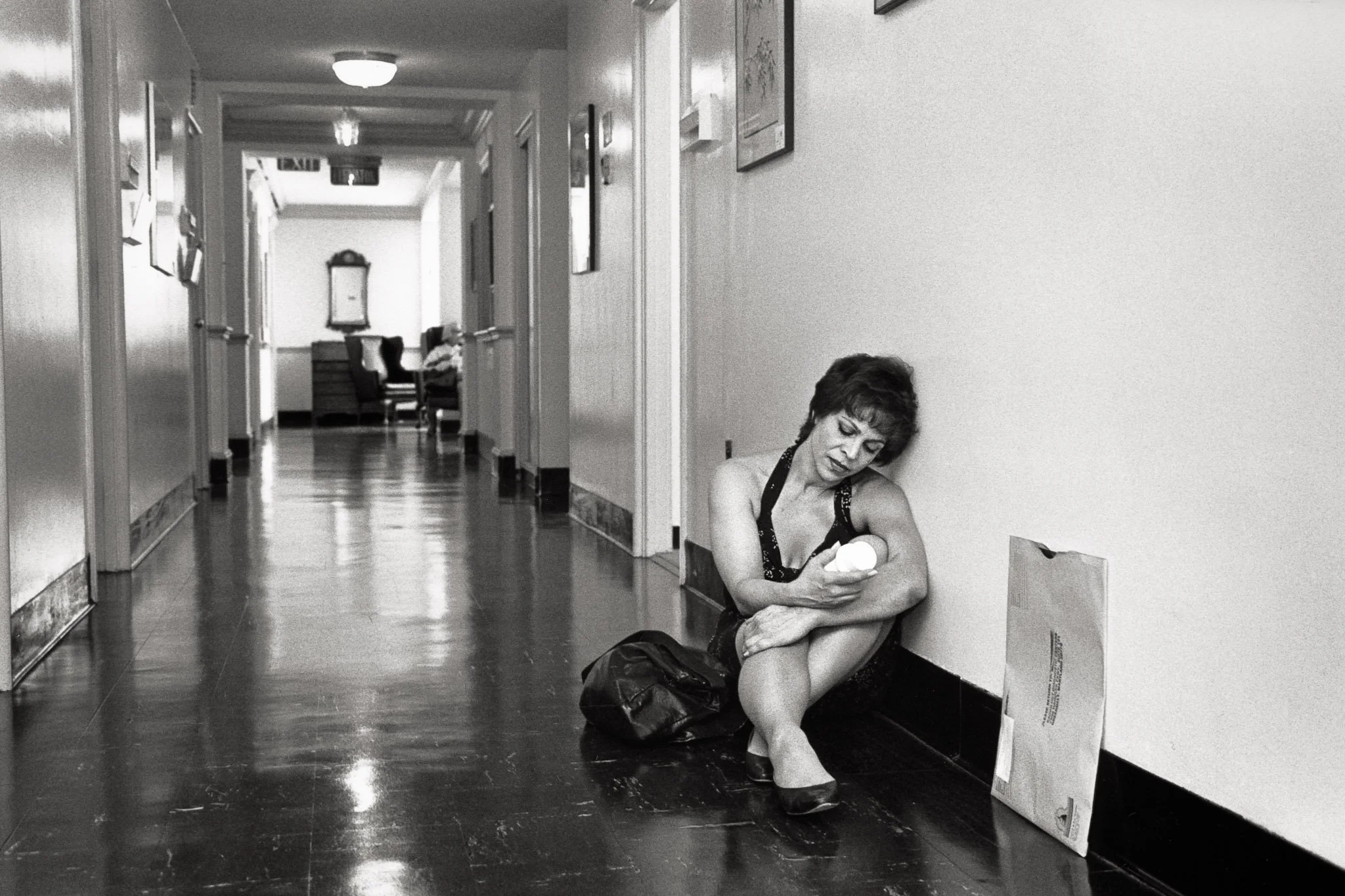 A woman in a dress sits on the floor of a medical building hallway, looking at a bottle of barium solution she must drink for a CT scan, a large folder with her medical scans next to her.