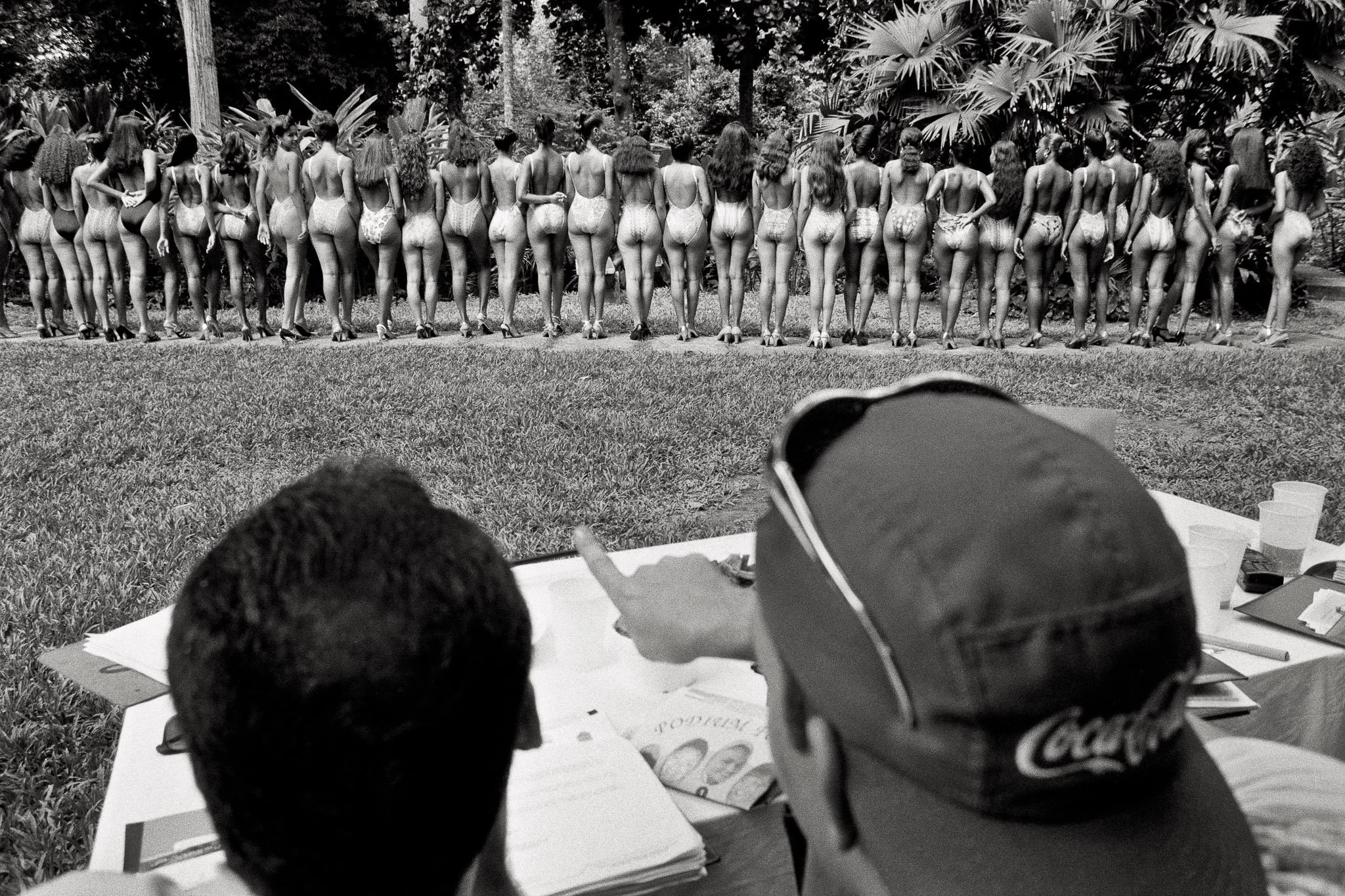 View over the shoulder of beauty pageant judges in Cartagena, Colombia. The contestants are lined up, facing away from the judges, so the judges and evaluate their butts. Image emphasizes the meat market feel of the pageants.