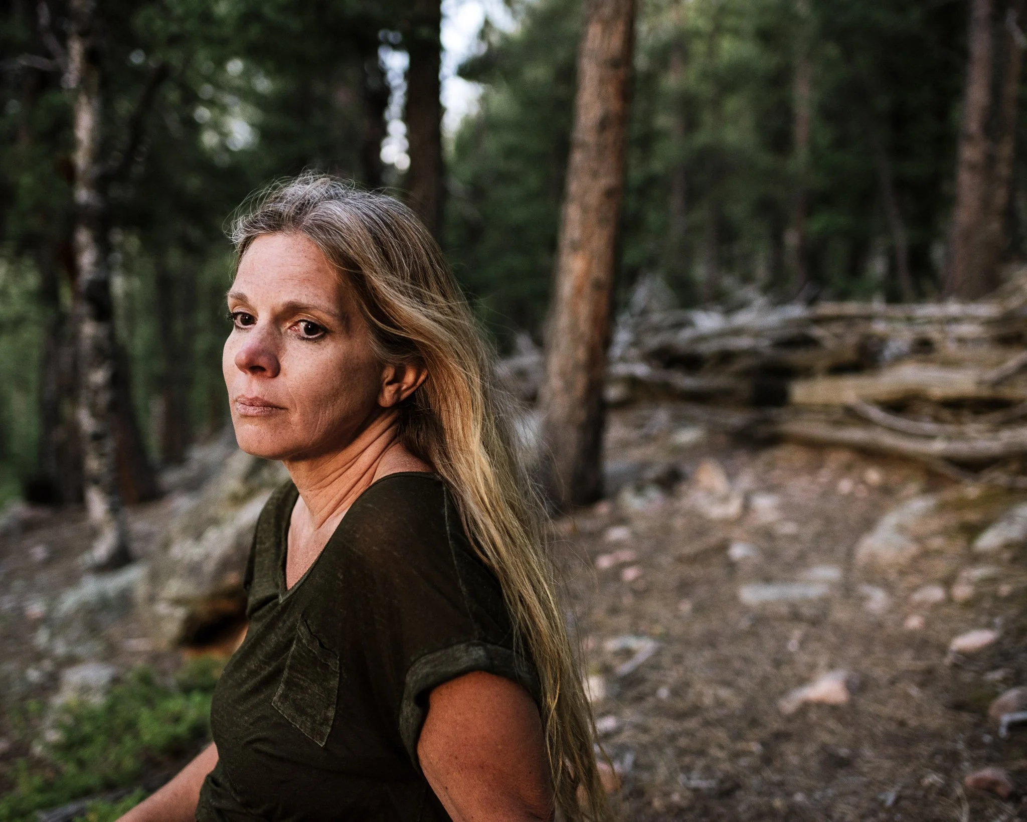 Woman with blonde/gray hair in a pine forest at dusk, lost in thought with a melancholy expression. She mourns the loss of her son.