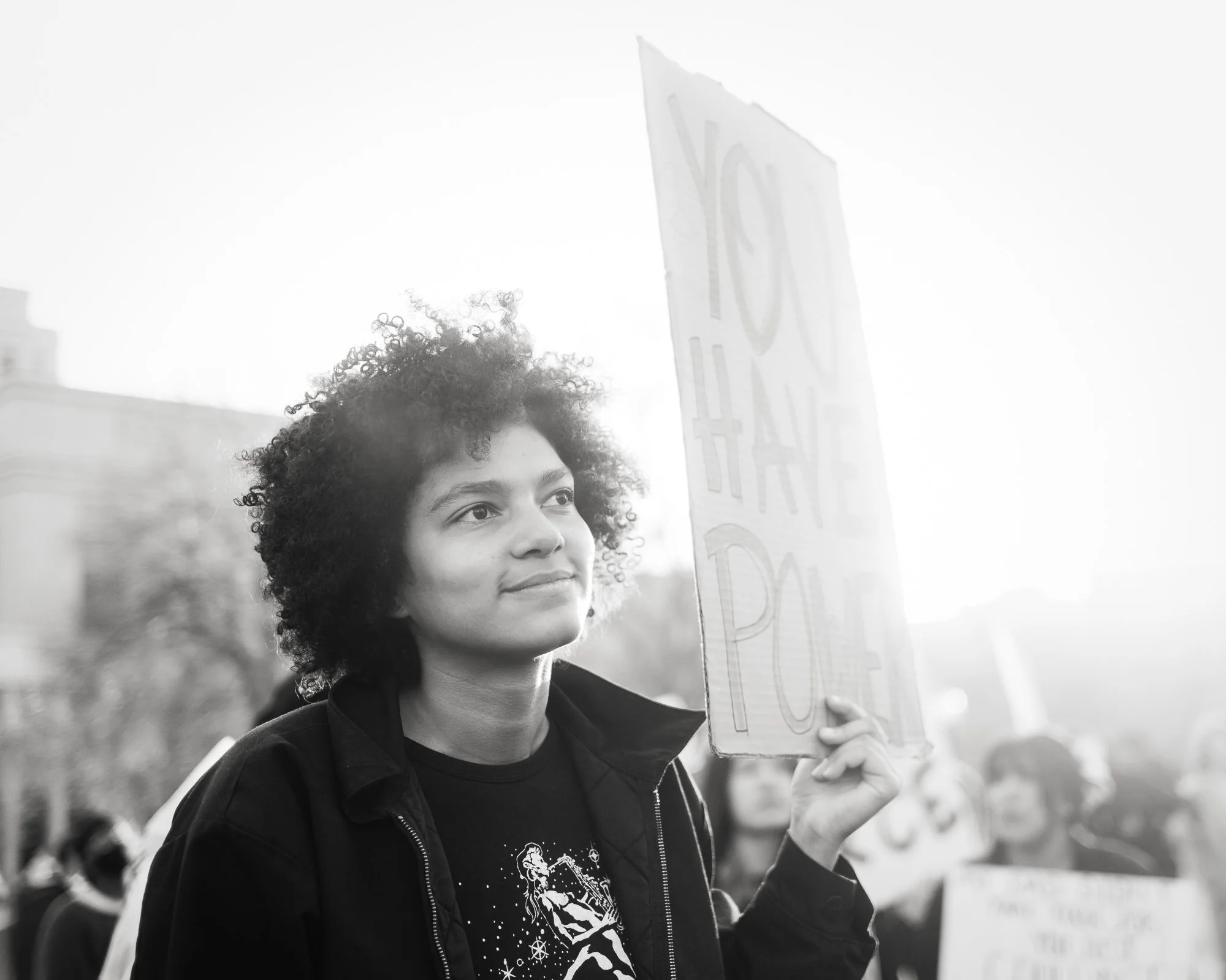Beautiful young black woman at a demonstration in the  late afternoon, gauzy iight, a subtle smile on her face, kind eyes, homemade sign reading "You Have Power"