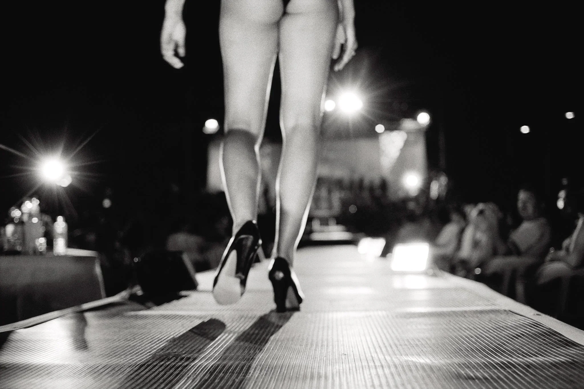 View of a woman on the catwalk of an outdoor pageant in Colombia, the cropping suggesting that she is naked aside from her high heels, walking into the lights and onlookers, highlighting the physical nature of the contests.