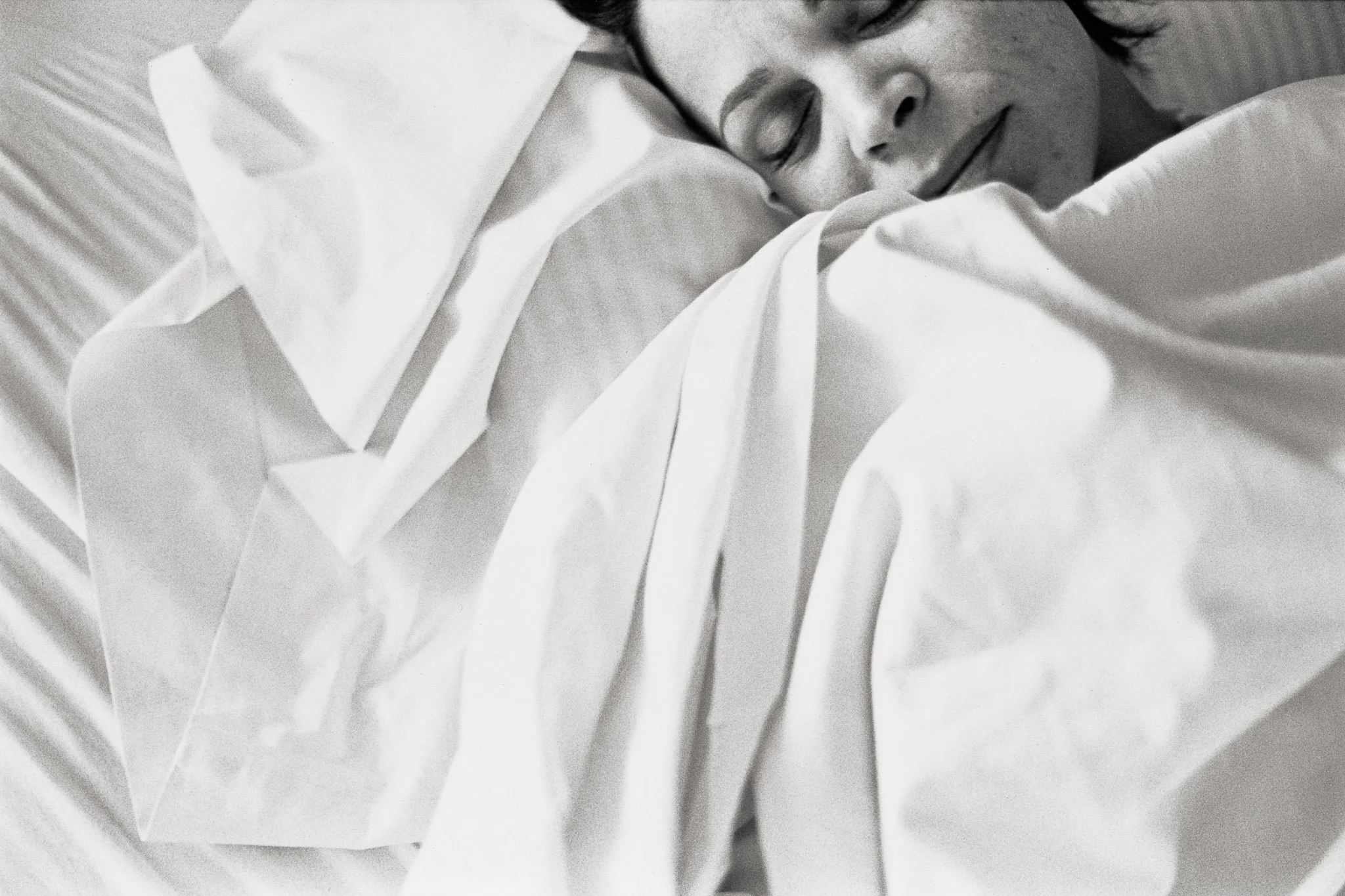 High key image of a woman sleeping in white sheets, the folds and creases almost ethereal.  Her face is just visible on edge of the frame, tightly cropped, sleeping with a serene expression. The mood is of airy transcendence.