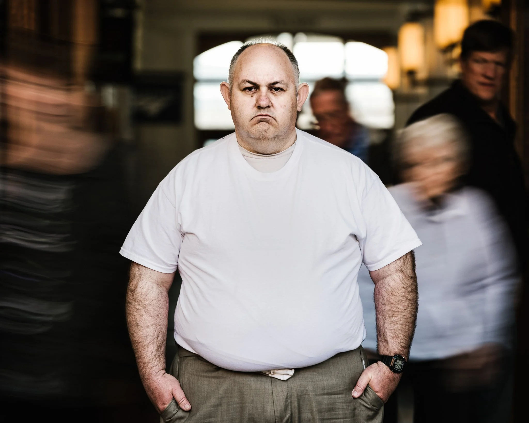 Portrait of a large man in a white t-shirt and a bitter expression, still as crowds swirl around him. He is afraid of never being loved.