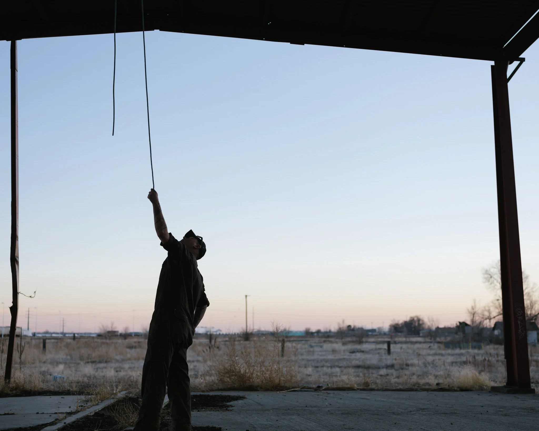 Silhouette of a young man in a large open abandoned factory after sunset in a high desert or prairie environment, testing ropes hanging from the ceiling.