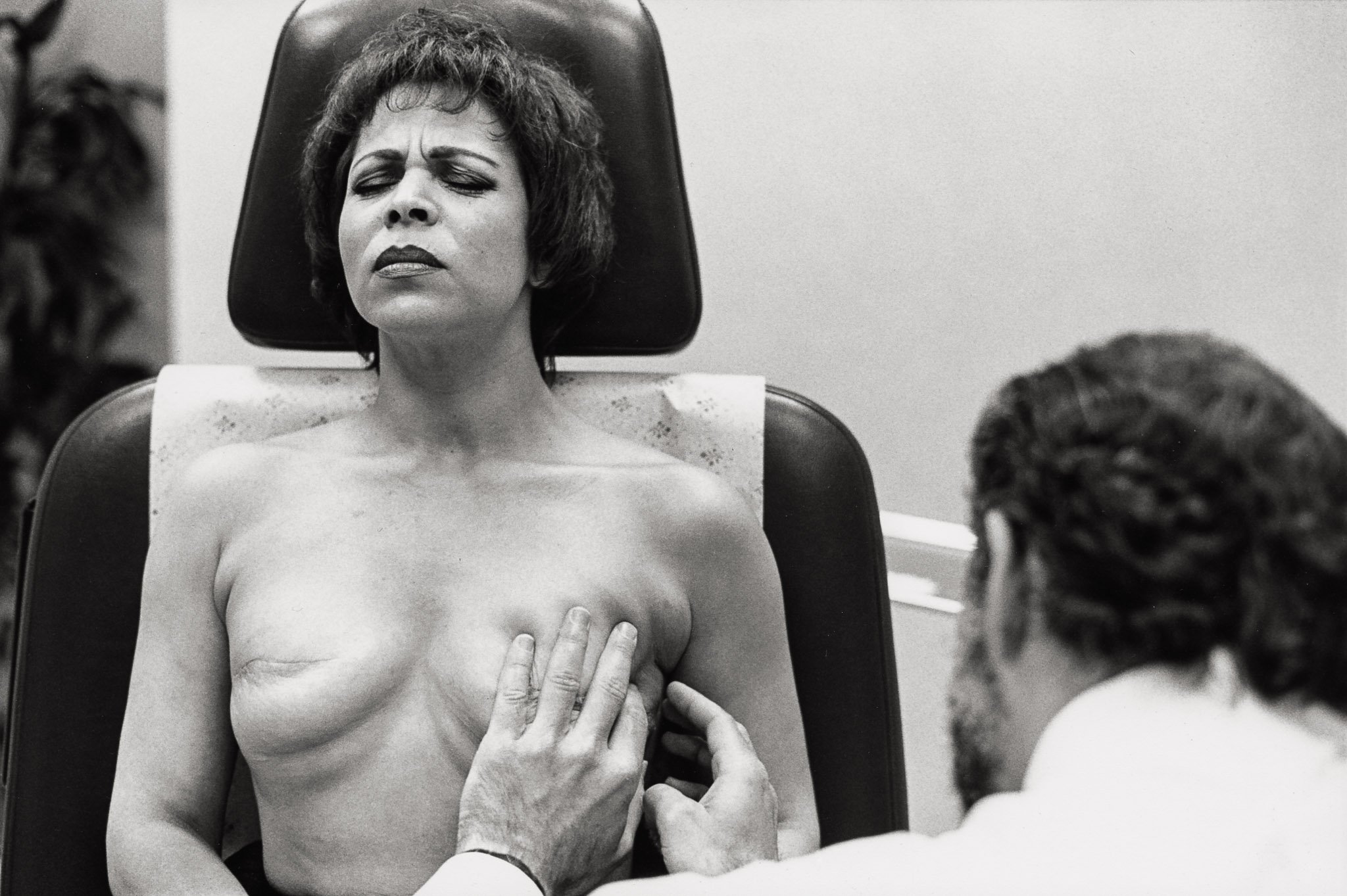 A woman with a bare chest sits against a chair in a plastic surgeon's office,  wincing as the doctor pokes at scar tissue after ruptured implants were replaced after a bilateral mastectomy. The pain is a relief, indicating her breasts are not numb.