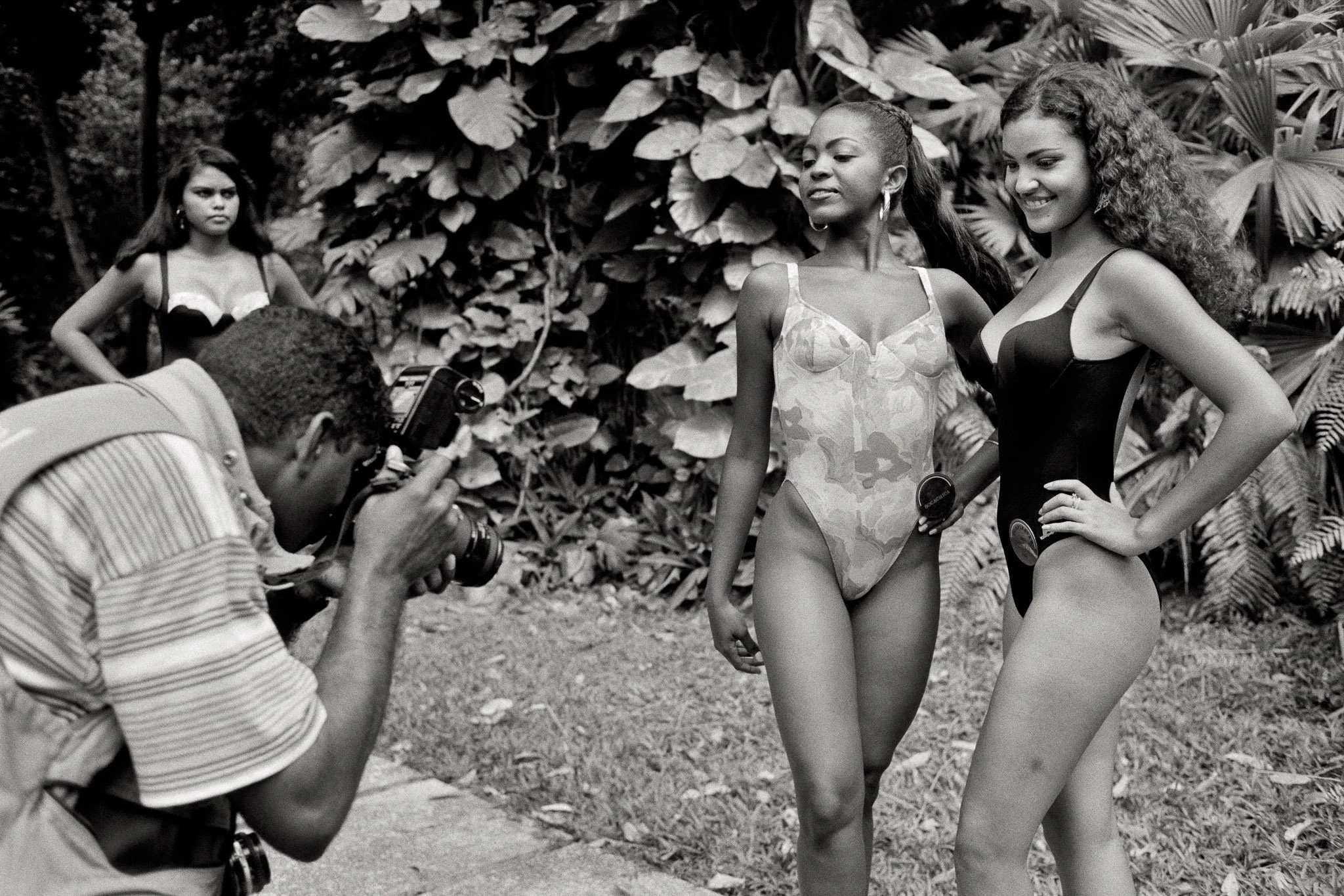 Two beauty queens pose for a local photographer near Cartagena, thick jungle foliage in the background. He is at close range, unmistakably taking pictures of their crotches. They smile. A third woman stands to the side with a critical expression.