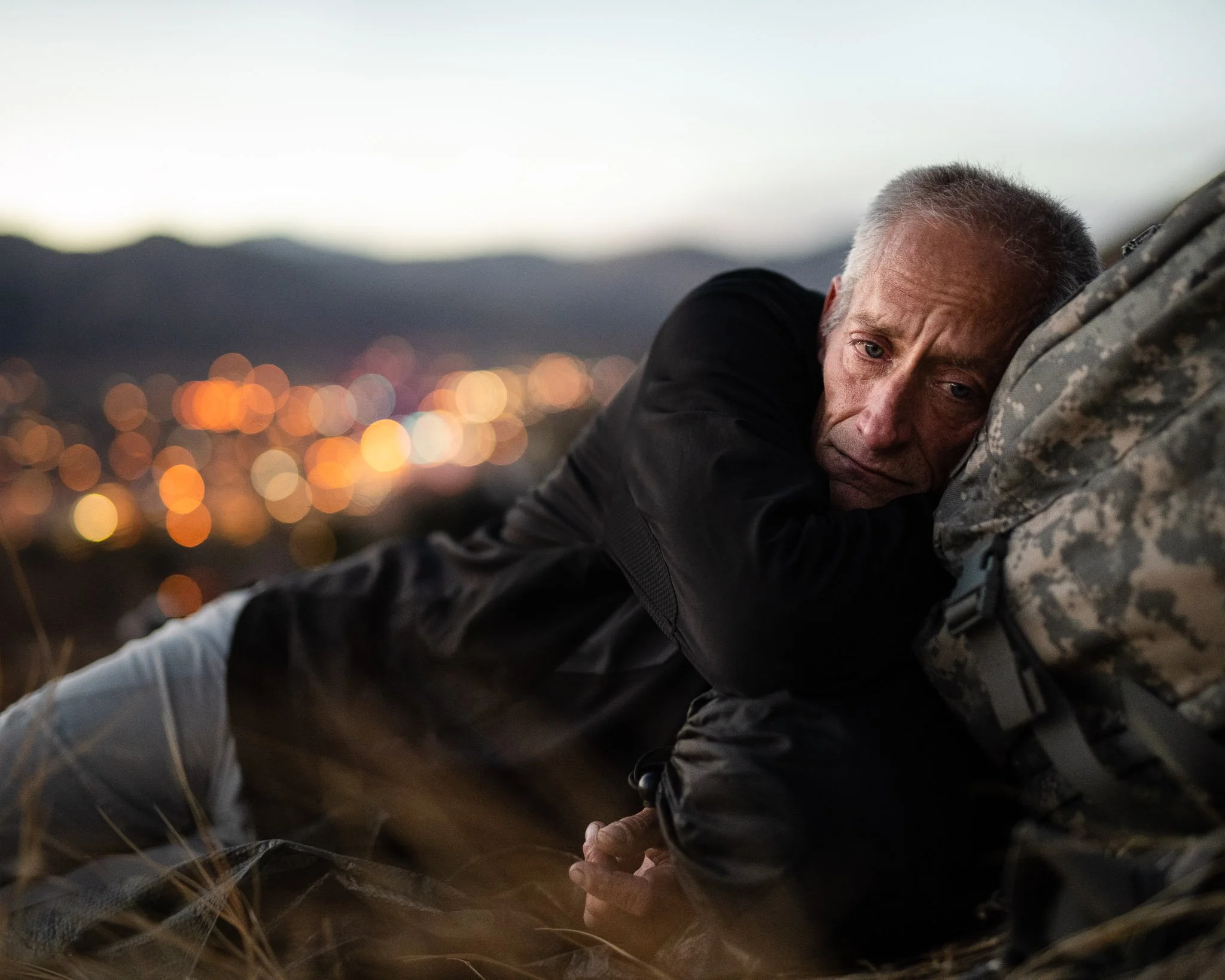 A middle-aged homeless man with short gray hair lies on the ground in the mountains at dusk in the cold, the lights of the city below him in the distance. He looks lonely, wounded.