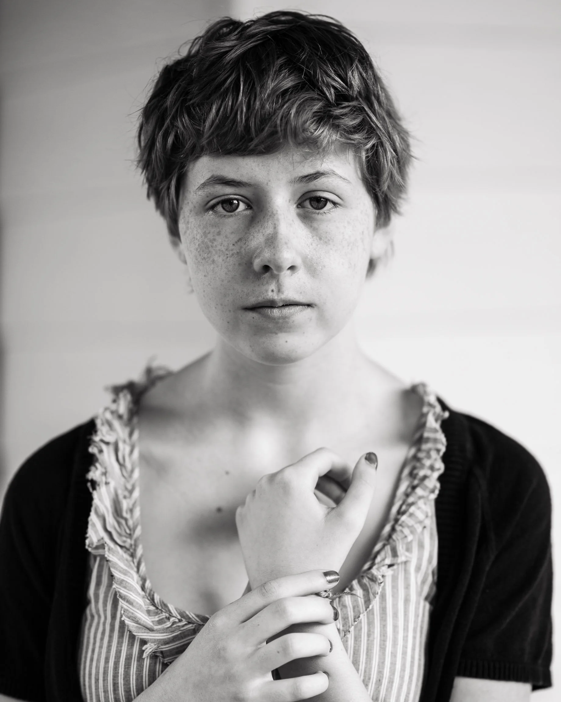 Monochrome head and shoulders portrait of a young woman with an expression both innocent and wise, her clothing simple and from another time, delicately holding her wrist, her hand drawn protectively to her chest.