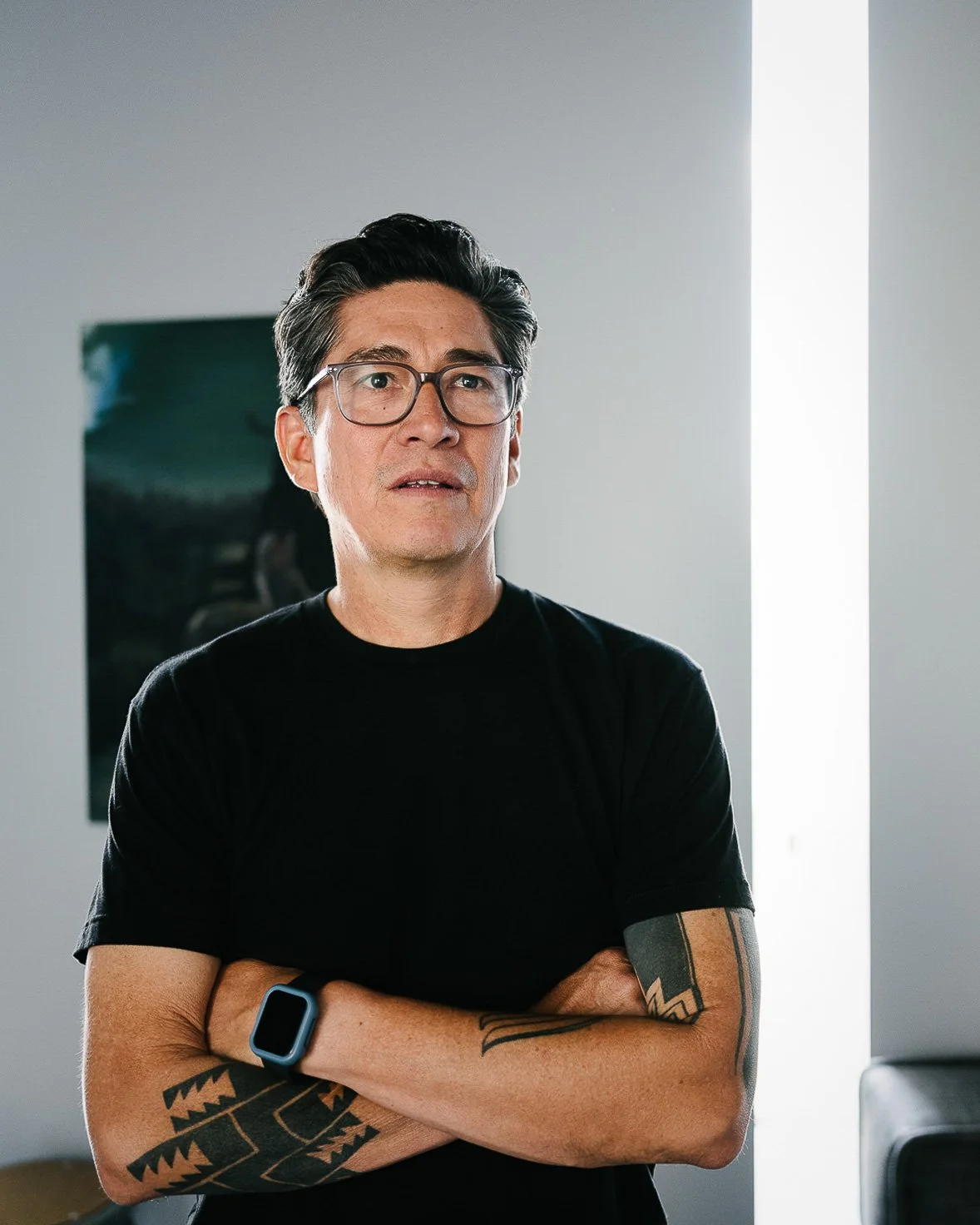Native American artist Steve Yazzie in a black t-shirt at work in his Denver Art Museum studio during his residency. He is evaluating a work in progress, his arms crossed, a thin bar of window light behind him.