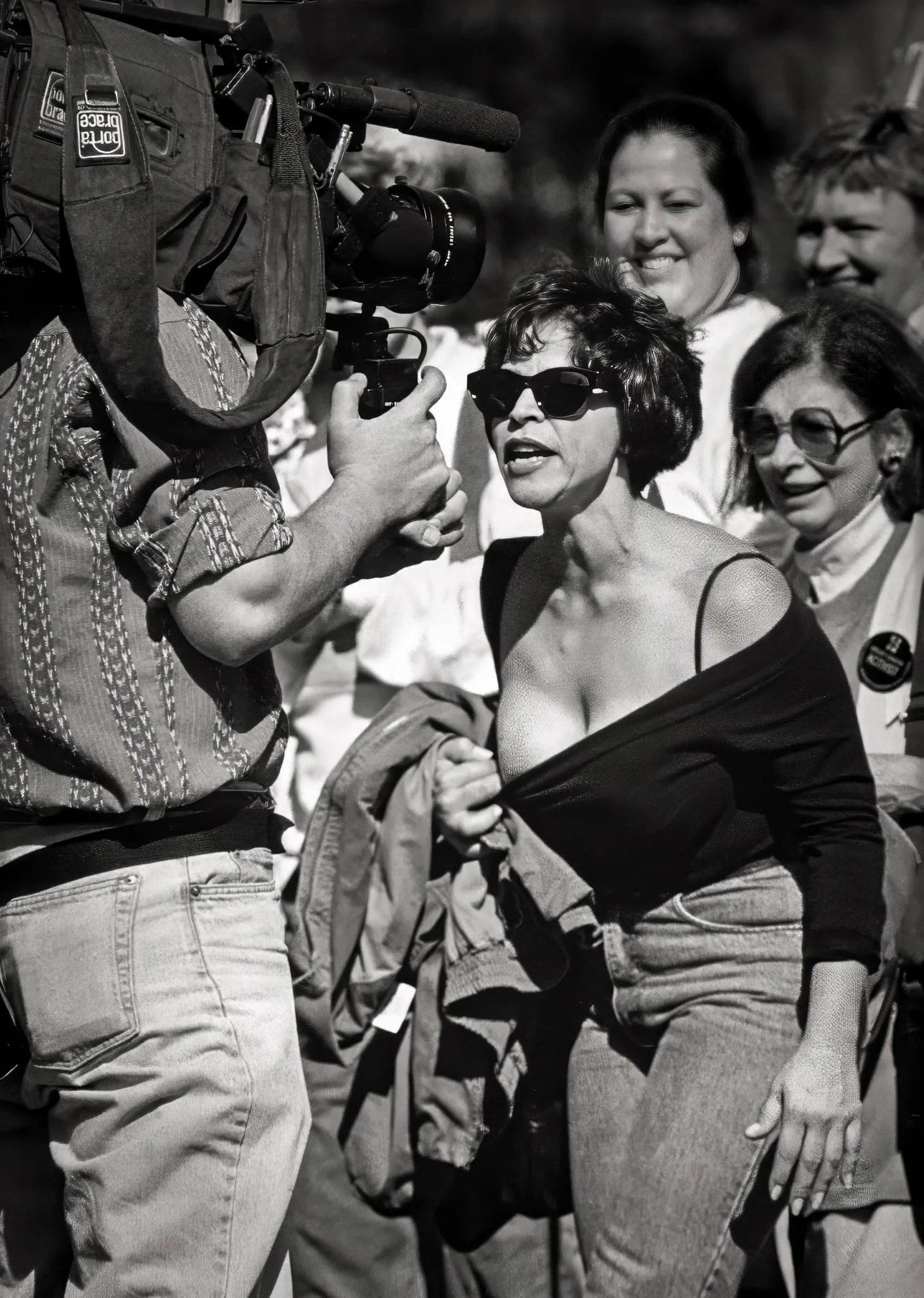 A woman at a demonstration pulls down her top in front of a television cameraman to reveal her partially reconstructed breast. She is angry, yelling at the cameraman, while the women in the background are smiling.