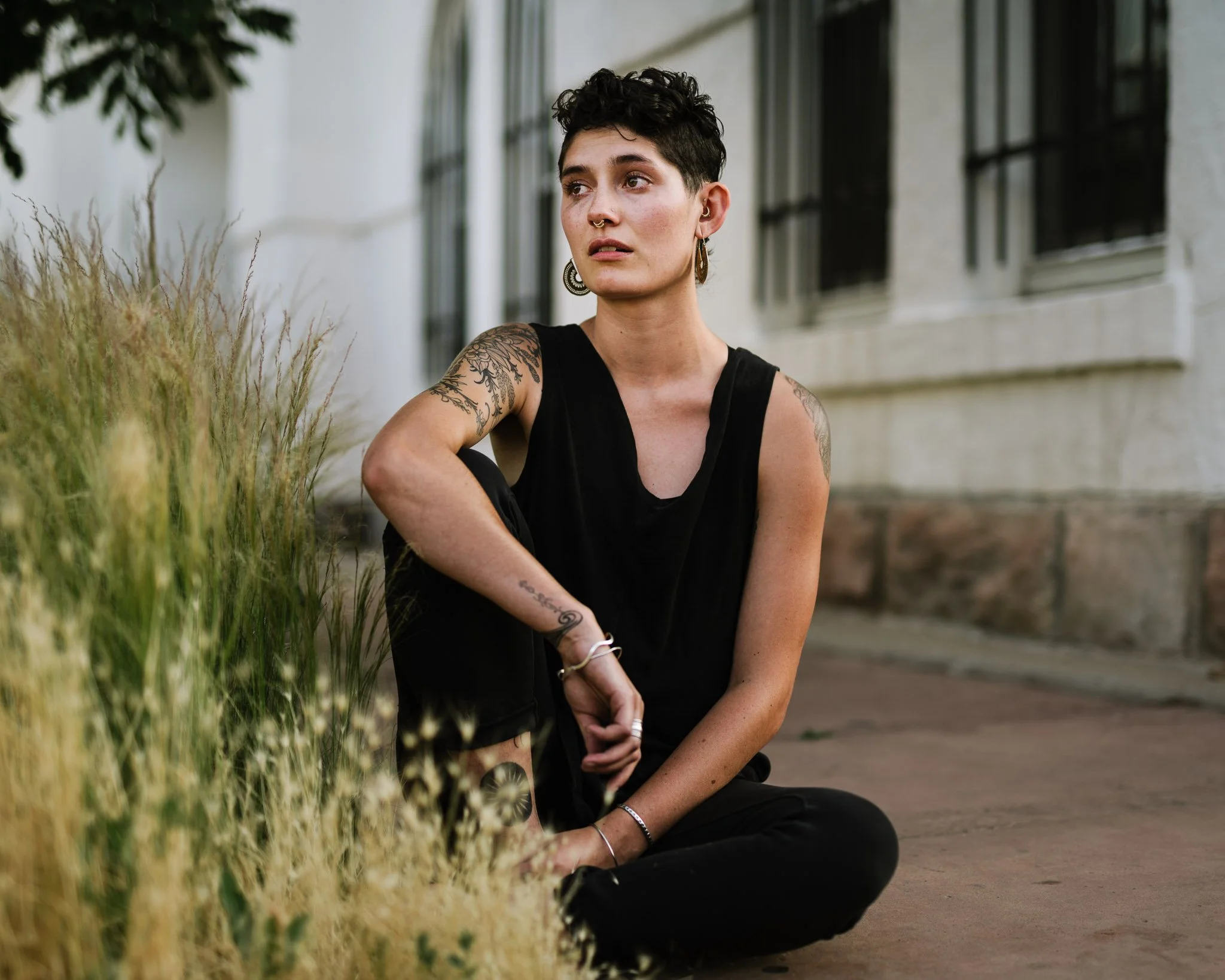 A young white woman in her late 20s sits on the sidewalk, next to tall grasses, dressed in black, her bare arms with tattoos and jewelry. She has short styled hair and large earrings, looking somewhat forelorn.