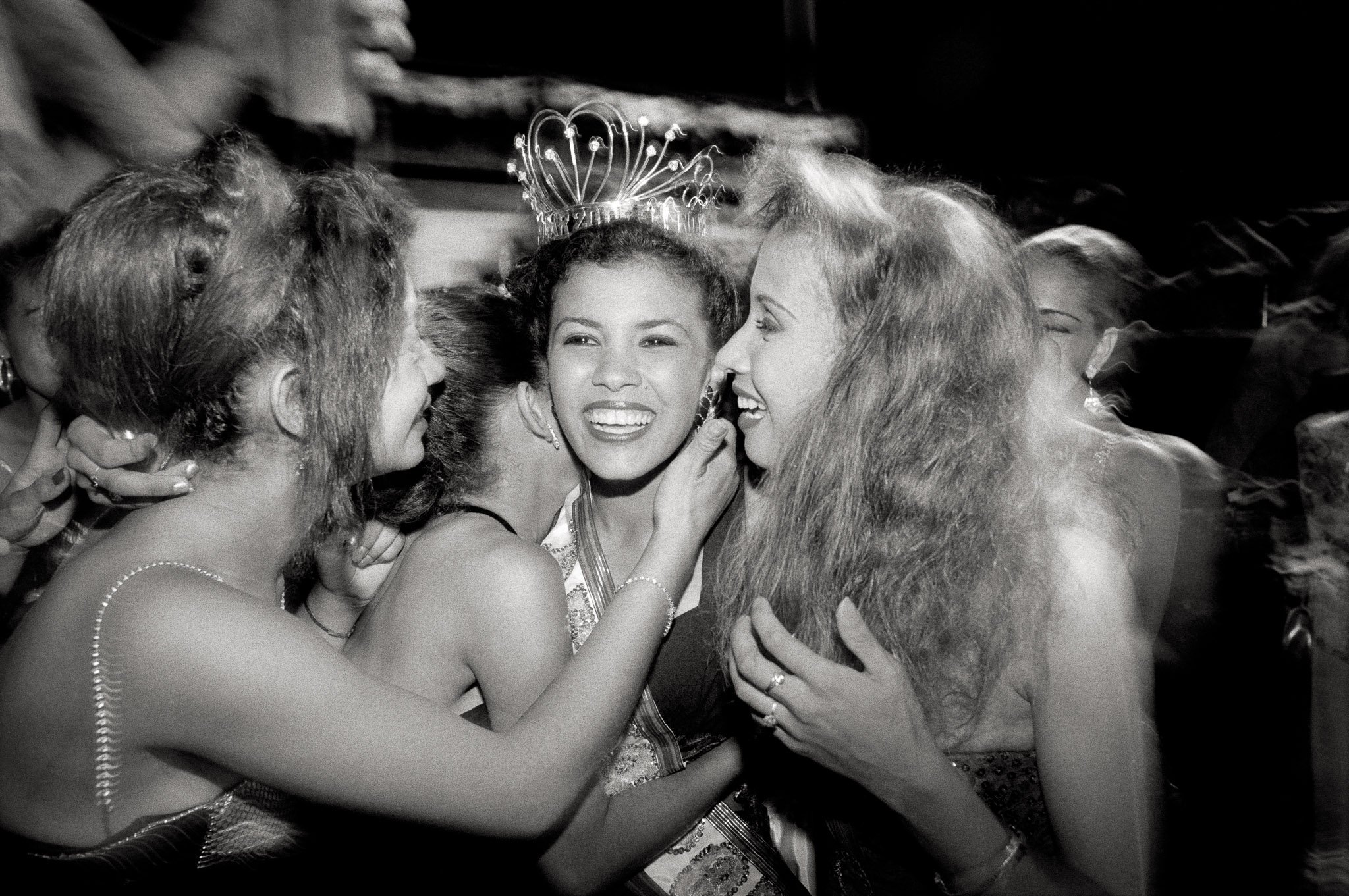 A young beauty queen with her sash and tiara beams while her fellow candidates embrace her. Close range picture, outdoors, with flash. There is an innocent exuberance and joy.