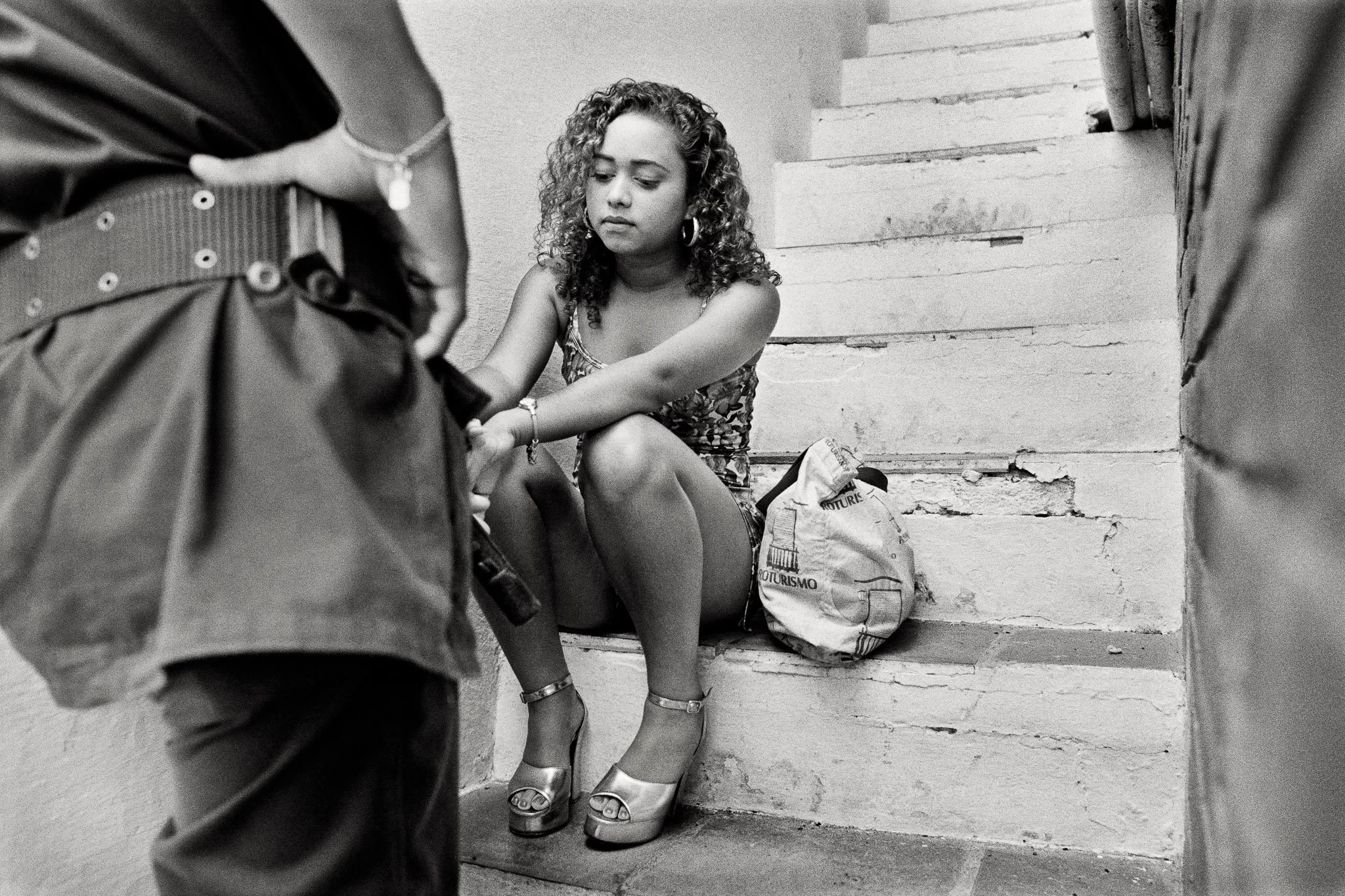 A beauty queen sits in a narrow stairway, high walls on each side, in a swimsuit and platform heels. A soldier stands over her, cornering her, in a dominant macho pose. She ignores him.