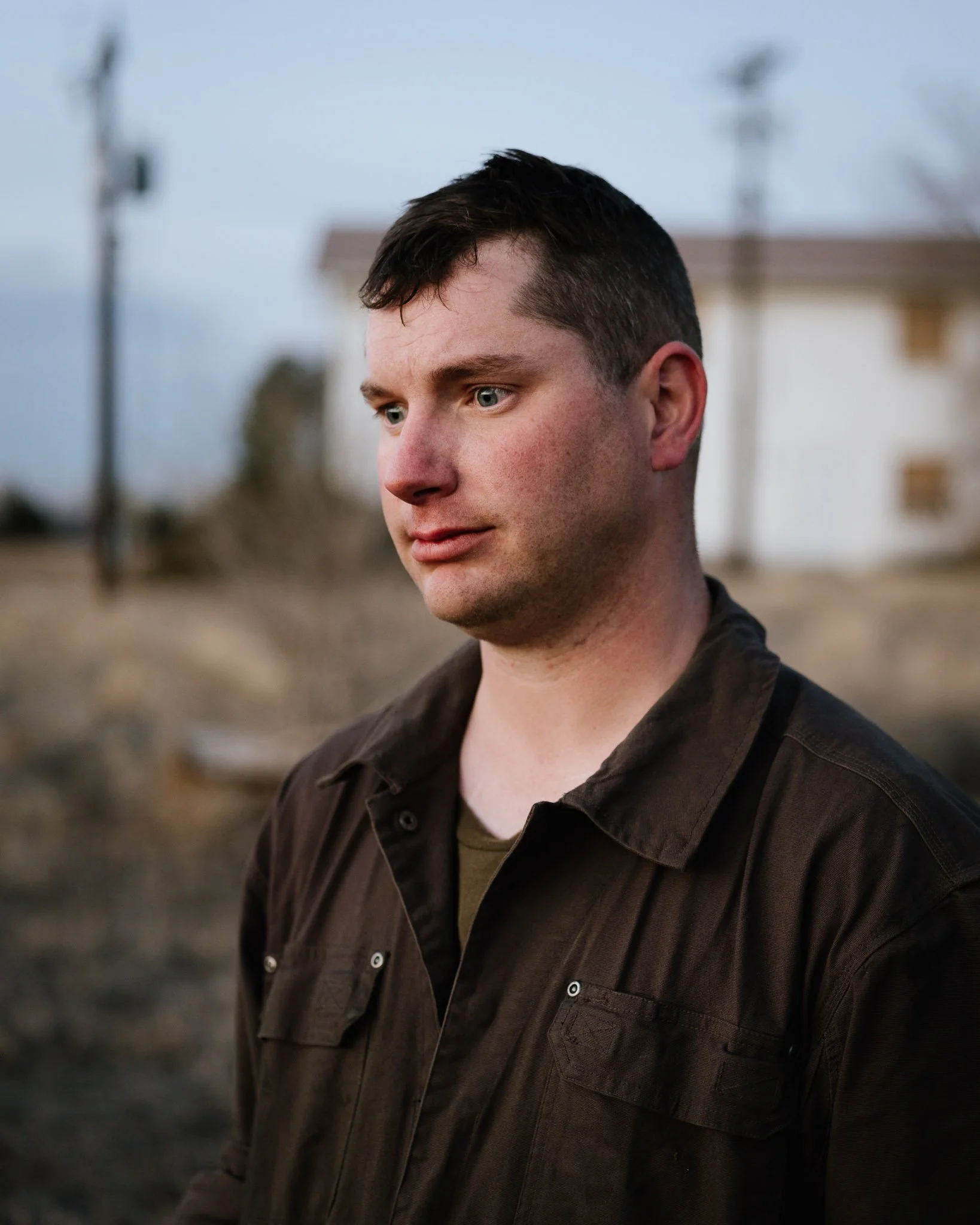 Photograph of young man in brown coveralls, a look of dismay on his face, ruddy complexion, faux hawk, at dusk near abandoned buildings.