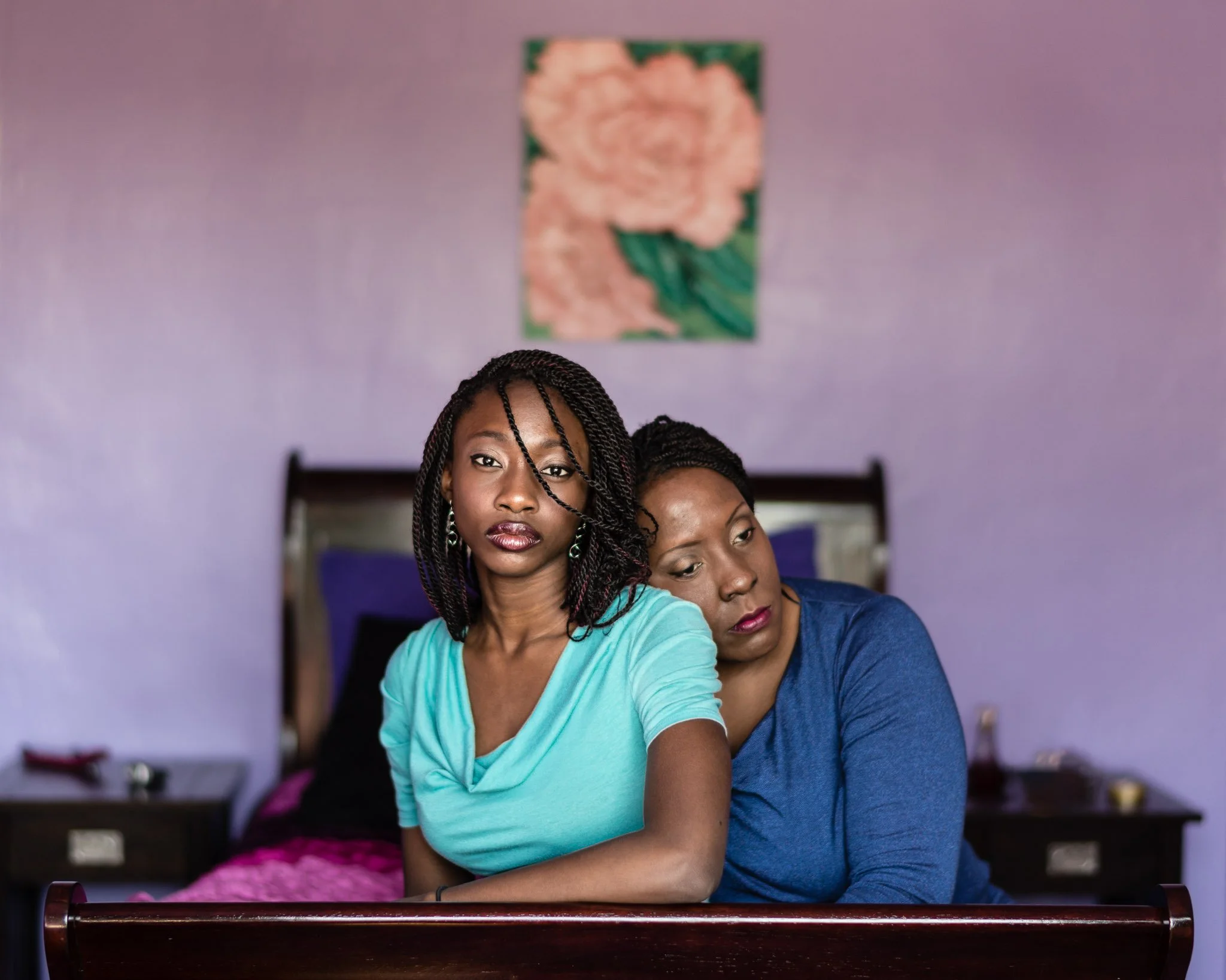 An adolescent black woman posing at the foot of her bed with her mother, her clothing, bedspread and the walls bright hues of blue, purple and pink. She gazes at camera, her mother behind her  with her head on the girl's shoulder, looking  away.
