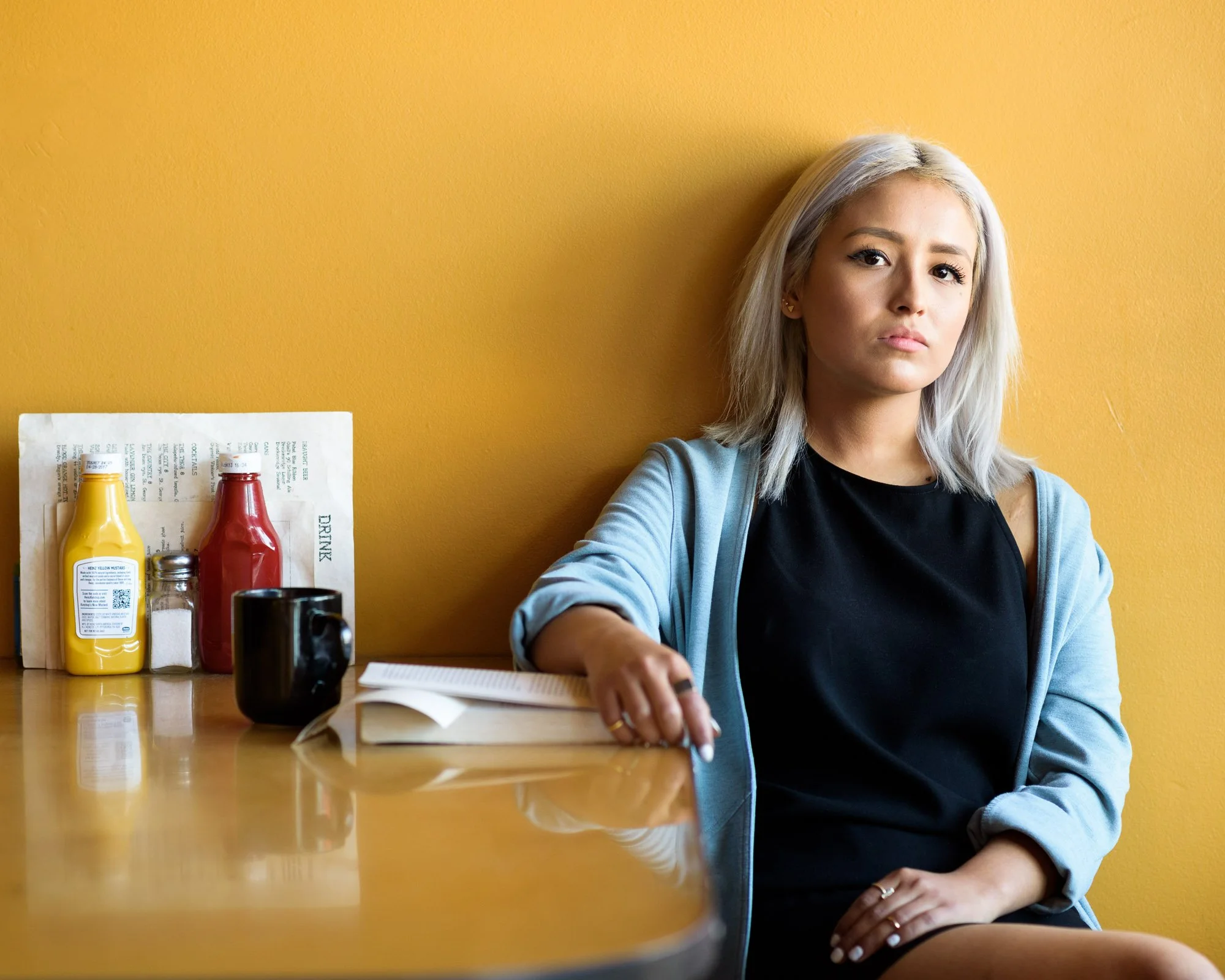 A light-skinned latina with platinum shoulder-length hair leaning against the yellow wall of a diner, her hand on a book on the yellow table, in a black sleeveless dress and blue sweater, her other hand on her thigh, looking at camera.