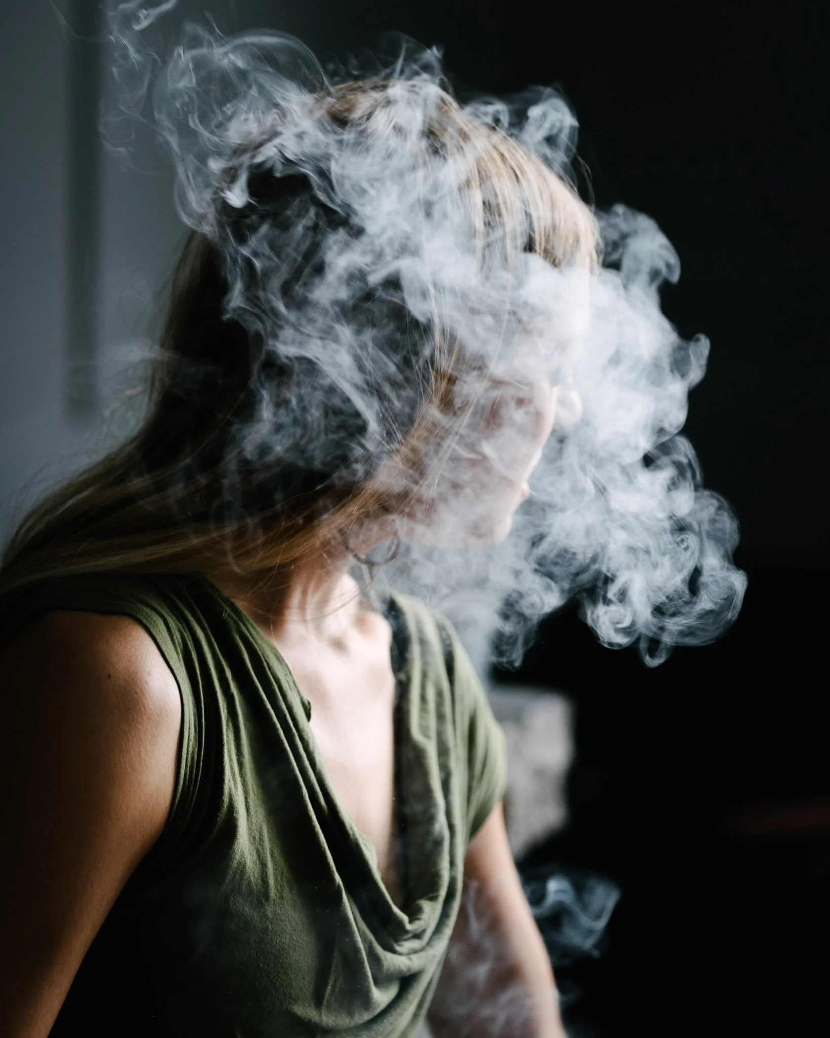 A woman with straight blond hair turned away from the camera, her head enveloped in a cloud of marijuana smoke that obscures her features.. Dramatic side/rear lighting accentuates her form and the smoke.
