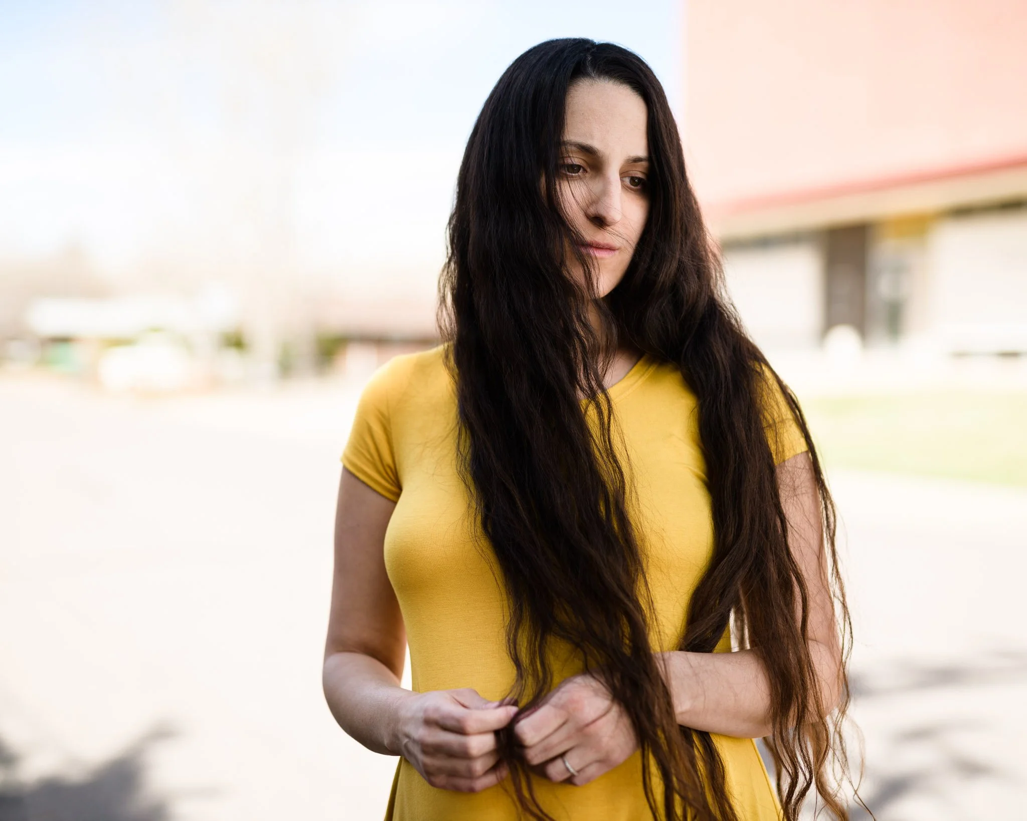 Woman with pale skin and long flowing hair and a yellow dress. She has a distant gaze reminiscent of a Renaissance painting stands before a washed out background. She is pregnant with dark circles under her eyes.