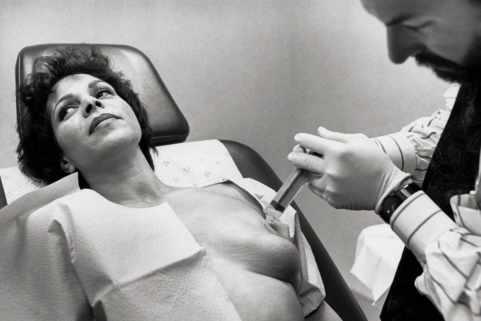 A woman leans back in a plastic surgeon's office as a large syringe filled with saline is injected into her implants for bilateral reconstruction after two mastectomies.