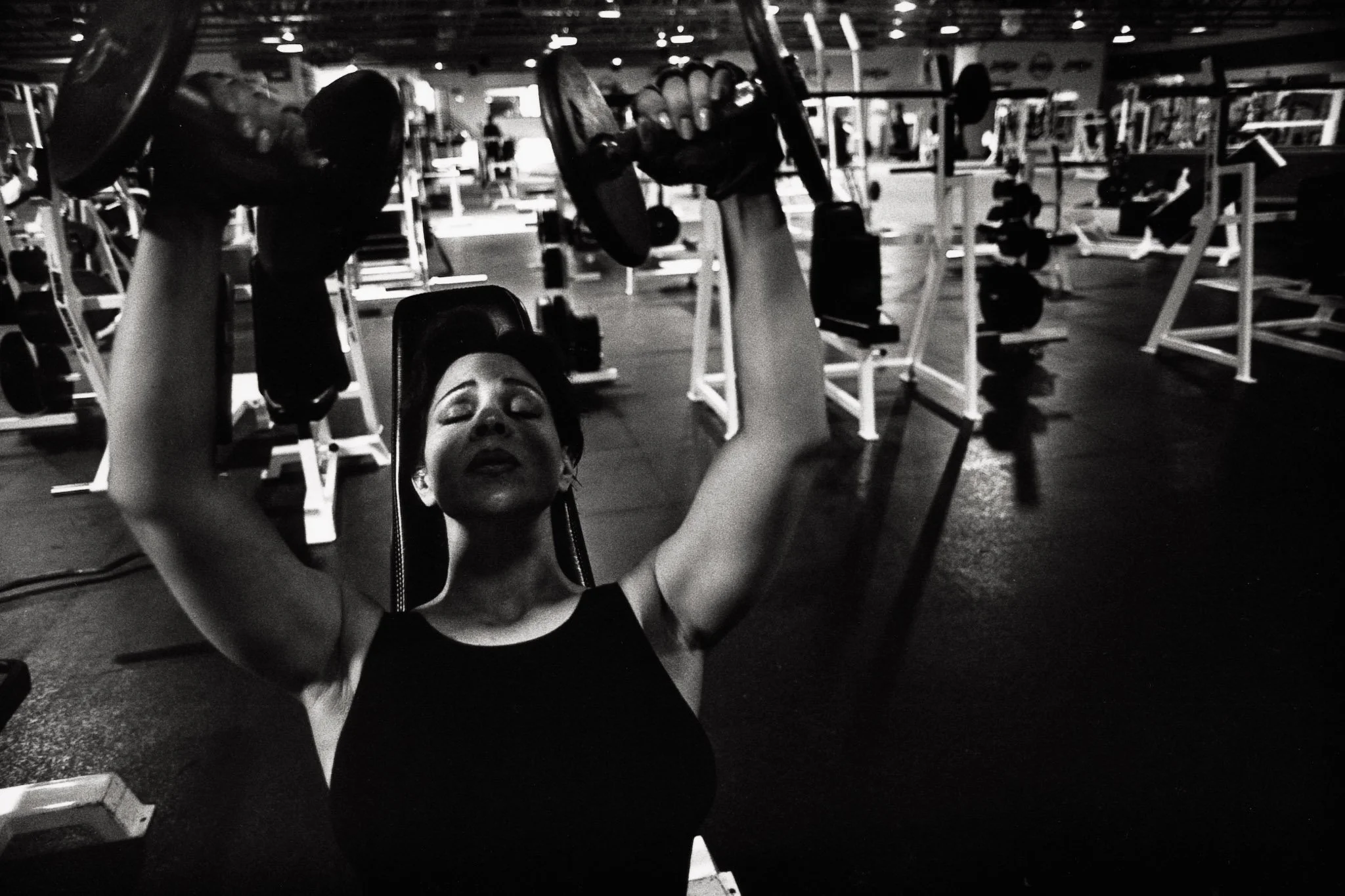 A dark frame taken in a gym, a woman on a bench lifts dumbbells over her head, eyes closed in concentration. She works to build her immune system to ward off a recurrence of breast cancer.