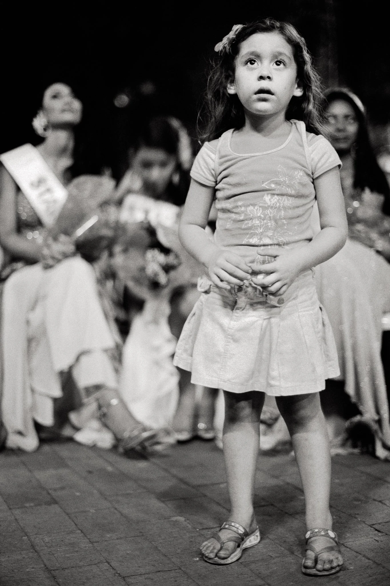 A young girl, perhaps four years old, in a t-shirt , skirt and sandals stares up at the stage of an outdoor Colombian pageant in awe of the queens on stage. Other queens are behind her in the background.