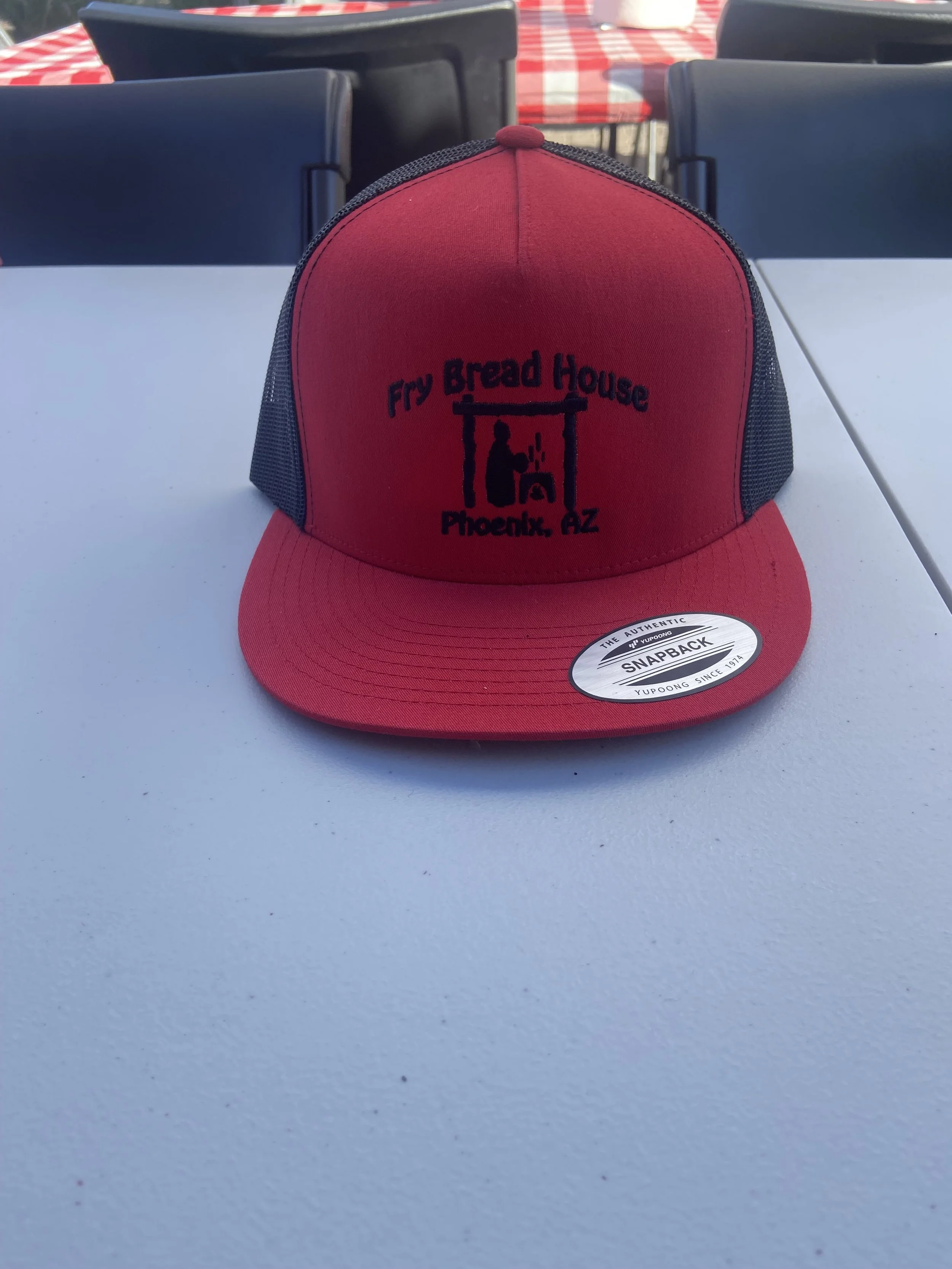 Red and black trucker hat with a logo for Fry Bread House in Phoenix, AZ, featuring a person baking bread in an outdoor oven.