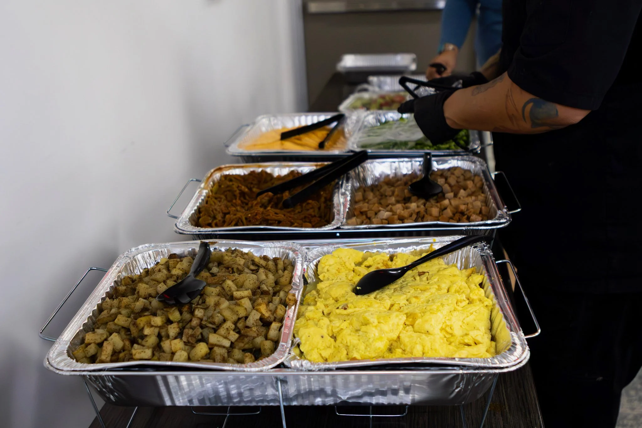 Buffet line with trays of scrambled eggs, diced potatoes, packaged meat, and vegetables, with people serving themselves.